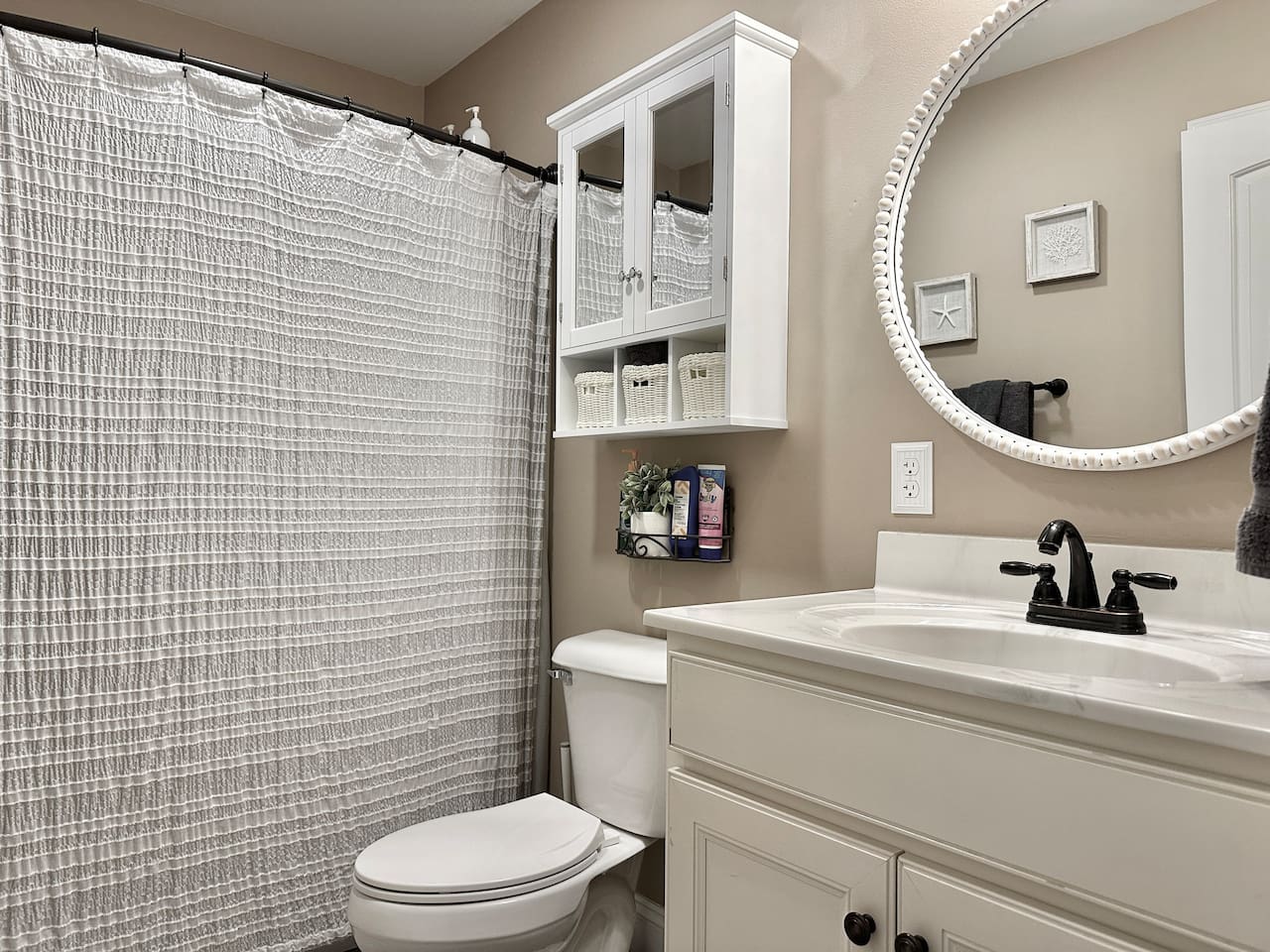 This bathroom features a white vanity with a dark faucet and a large, beaded-frame round mirror. It includes a white wall cabinet with glass doors, a standard toilet, and a shower area with a textured grey curtain.