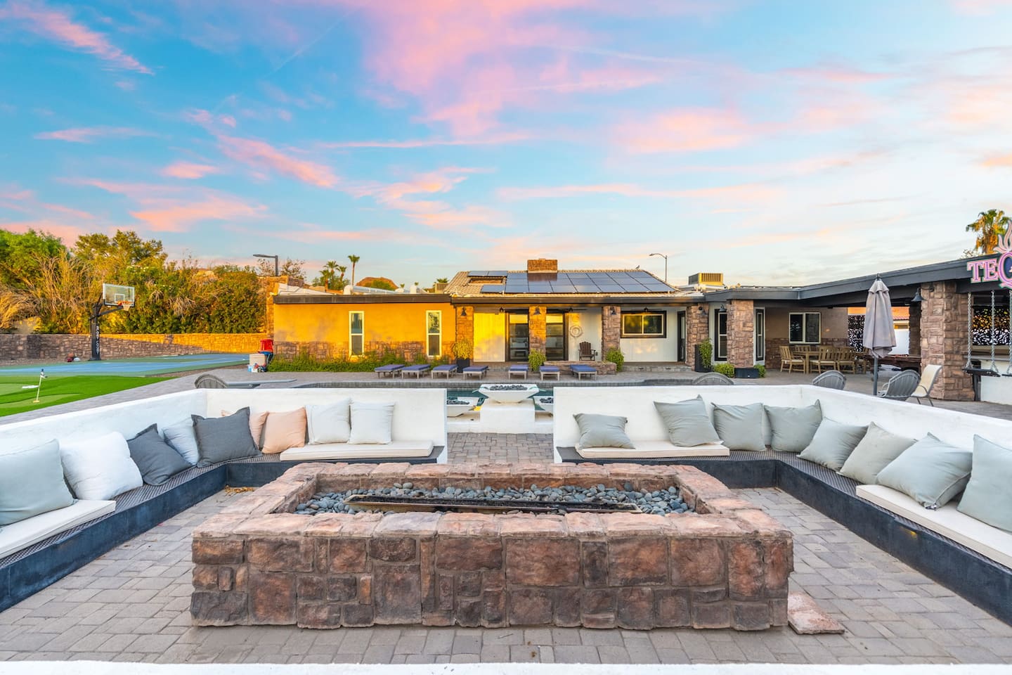 A built-in fire pit lounge with wraparound seating overlooks the pool, putting green, and sport court at dusk.