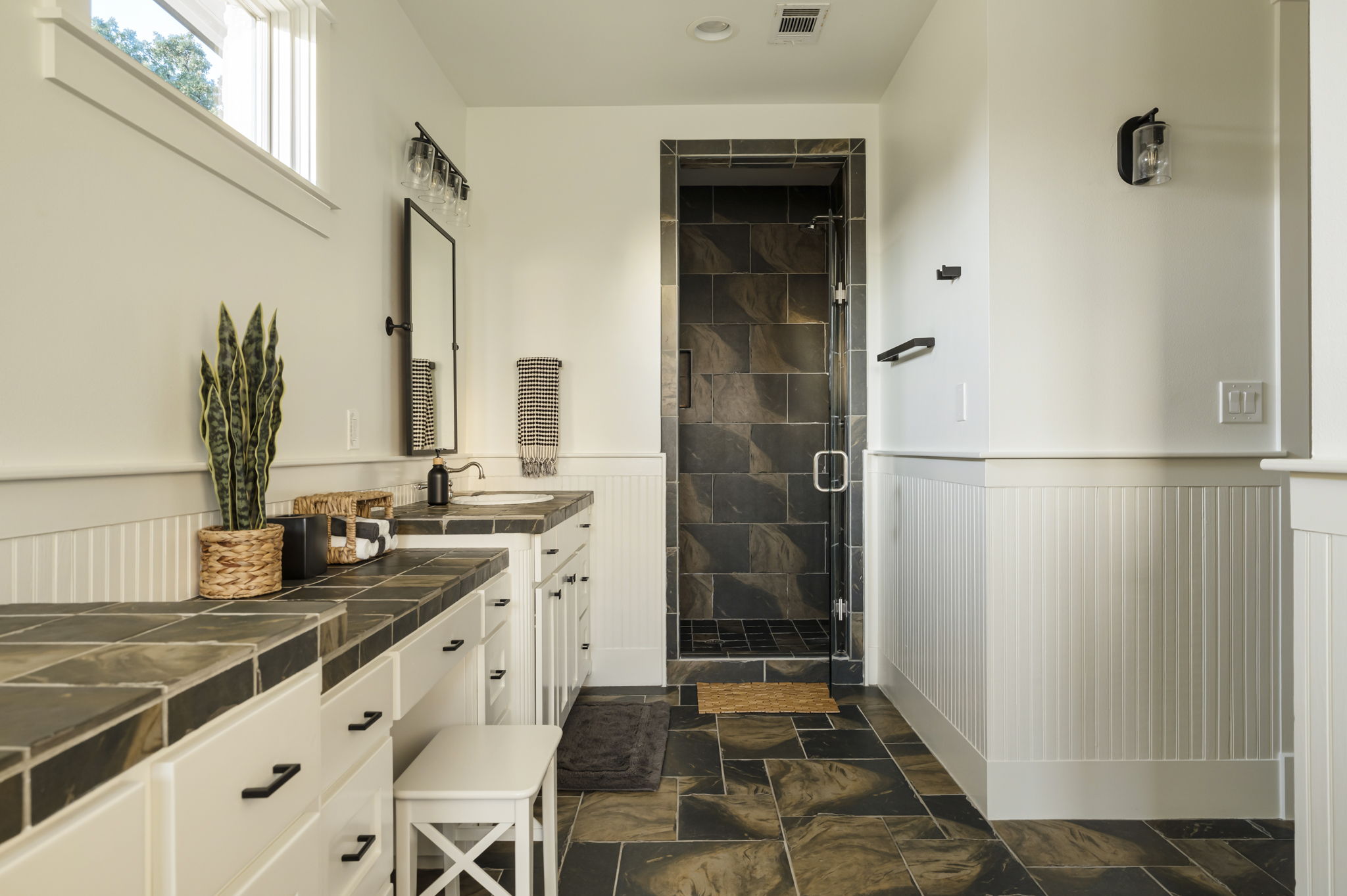 A walk-in shower with dark tiled walls and floor, creating a striking contrast with the bright white vanity area.
