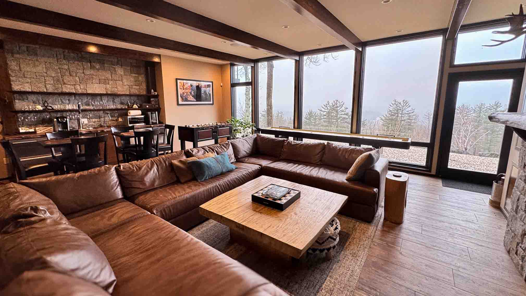The Tavern Room at Sunset Falls &mdash; leather sectional, live-edge bar, foosball table, and floor-to-ceiling glass to the foggy Berkshires ridgeline