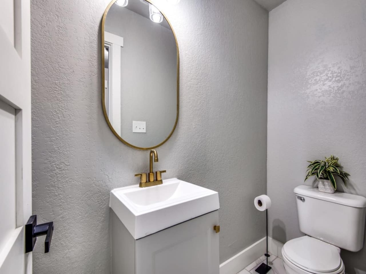 Stylish powder room off the kitchen with white pedestal sink, gold fixtures, and oval mirror. Added convenience for groups up to 7 in this 3BR/2.5BA modern farmhouse Airbnb in historic downtown Aubrey TX.