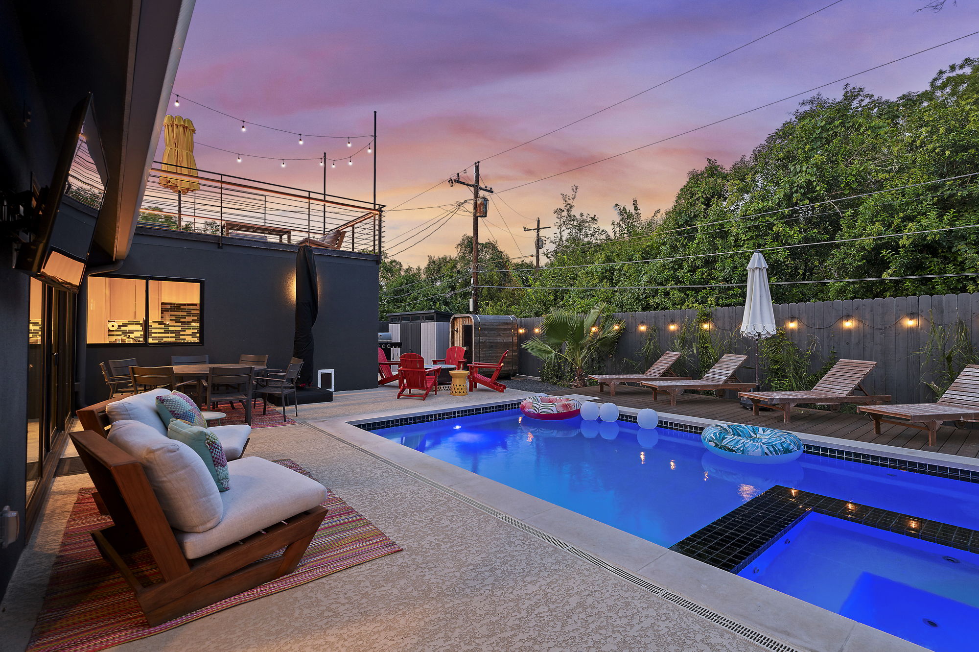 A vibrant backyard oasis at dusk, featuring a sparkling blue swimming pool, multiple lounging and dining areas with string lights, and a second-story deck, all enclosed by a modern wooden fence.