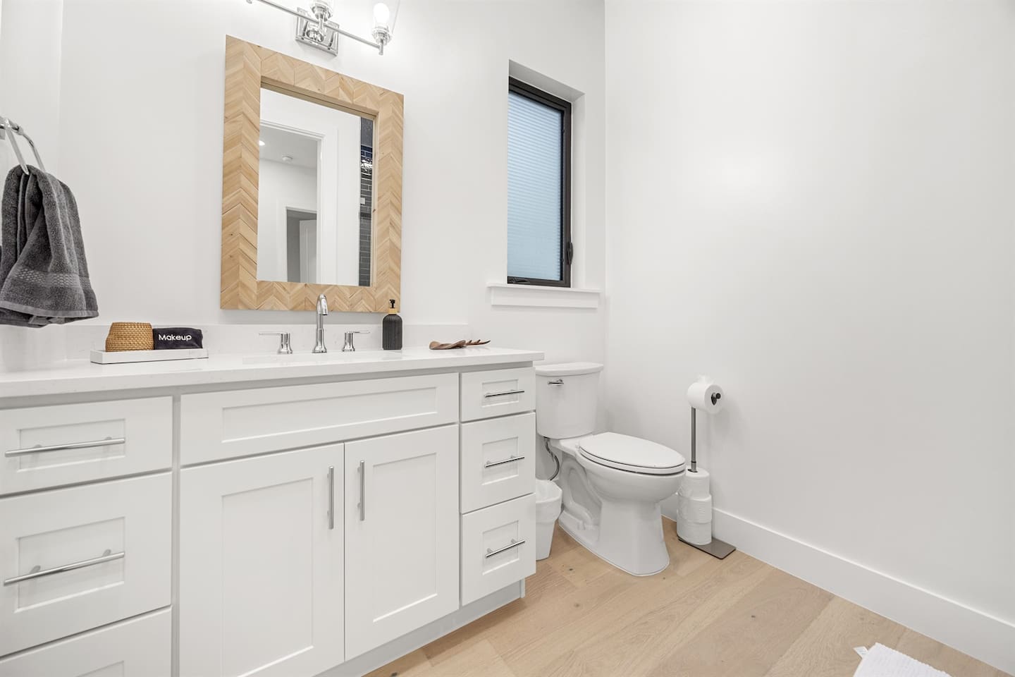 Discover a bright bathroom with a toilet under a window, a stylish sink and faucet atop a drawer, a wide designer mirror, neatly placed towel, and elegant lighting creating a refreshing ambiance.