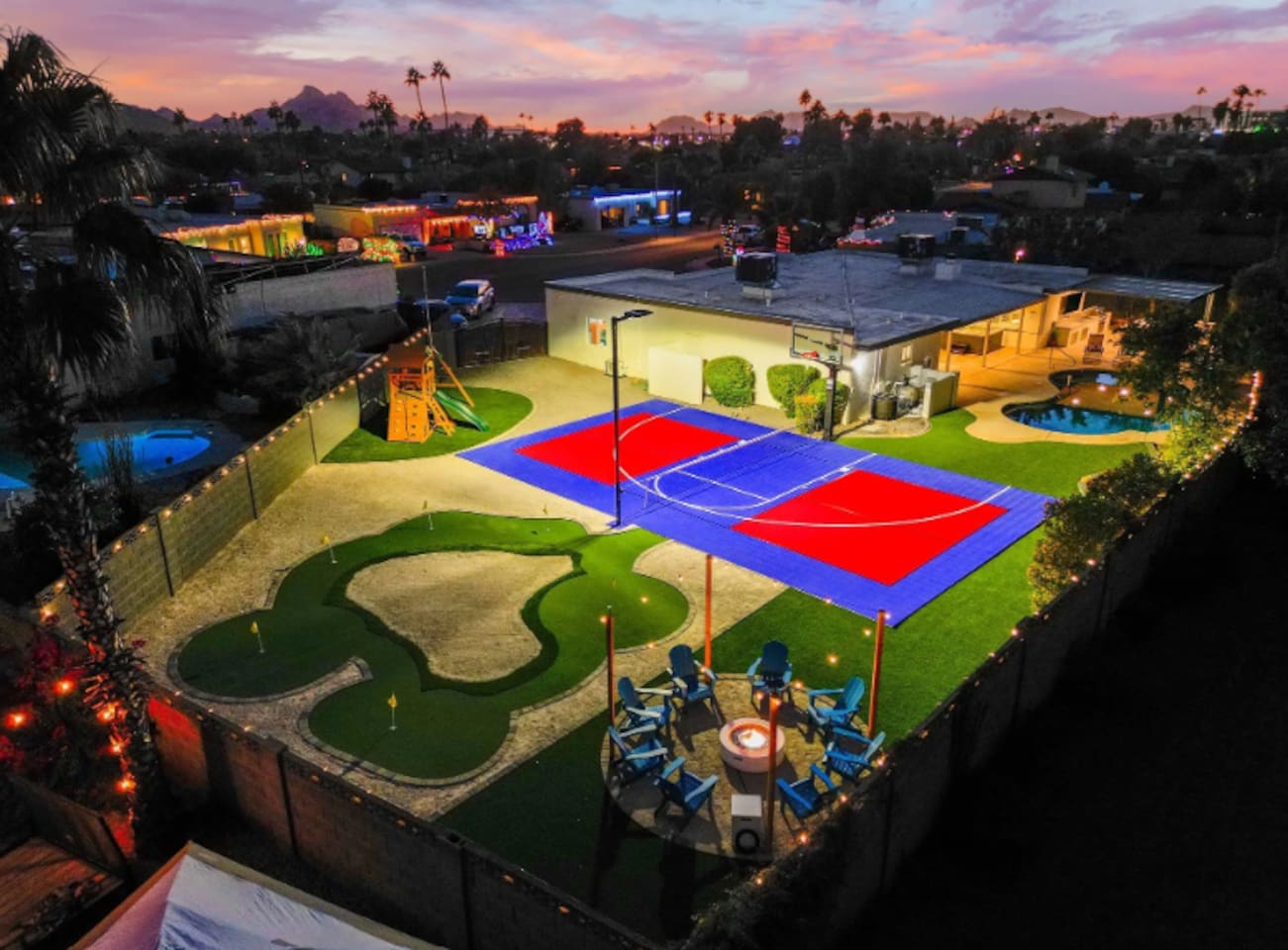 As the lights come on, the whole backyard turns into one big invitation — court, green, fire pit lounge, and string lights overhead.