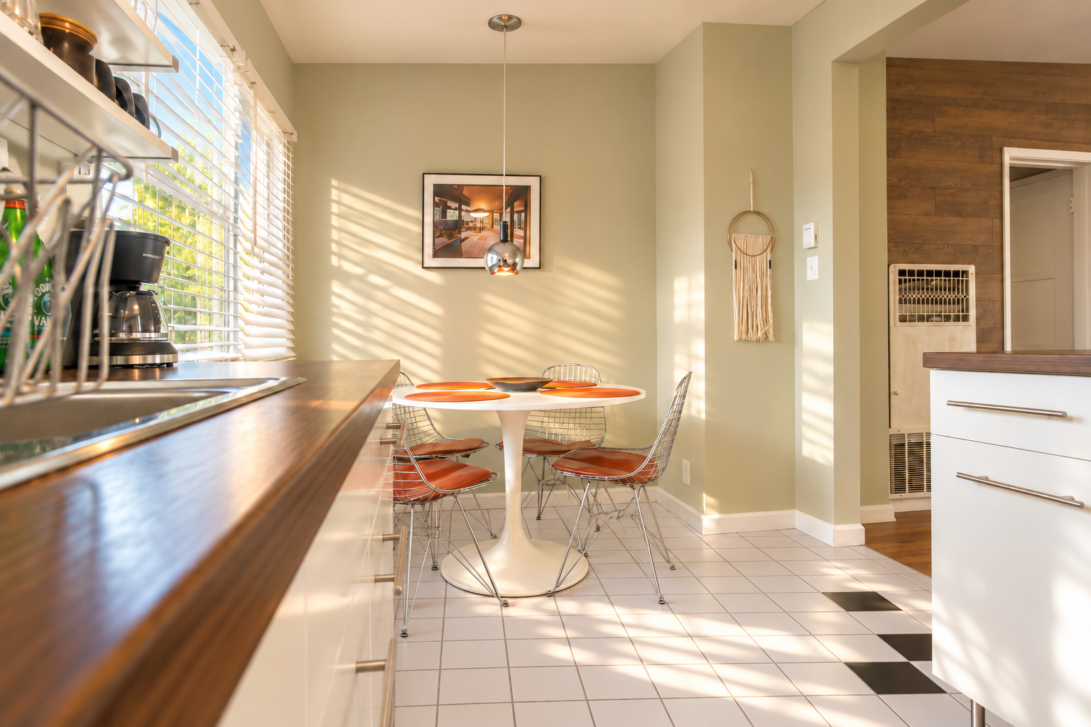 A sunny view along the wood kitchen countertop toward the round dining table and wire chairs. White cabinetry, a stainless-steel sink and pale floor tiles with dark accents keep the space bright, while the window brings in leafy outdoor views.
