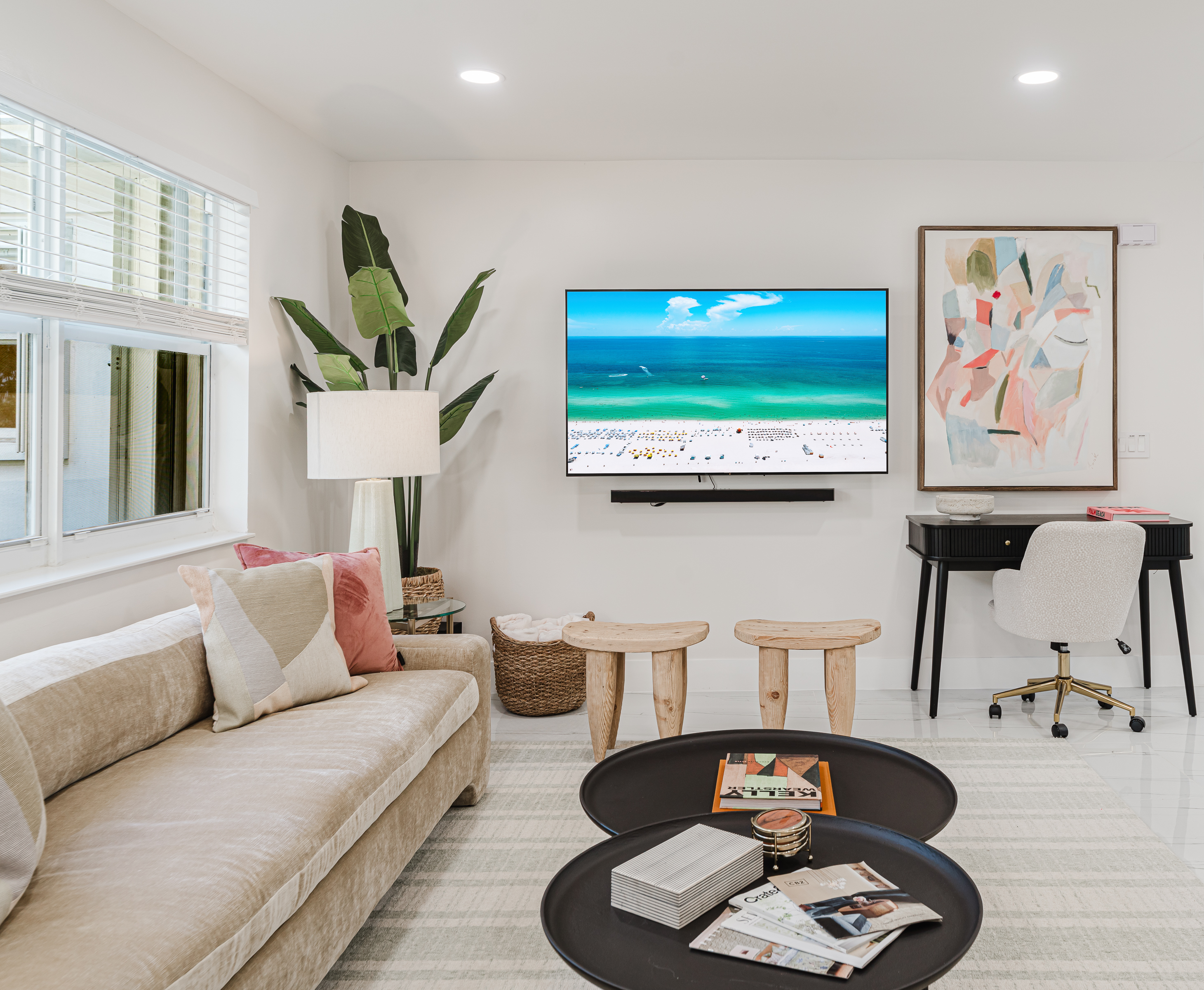 Light-filled living room with a 65" smart TV, sound bar, and subwoofer, featuring plush designer seating, neighborhood views, curated games, and reading materials—perfect for relaxing, entertaining, and family movie nights in West Palm Beach.
