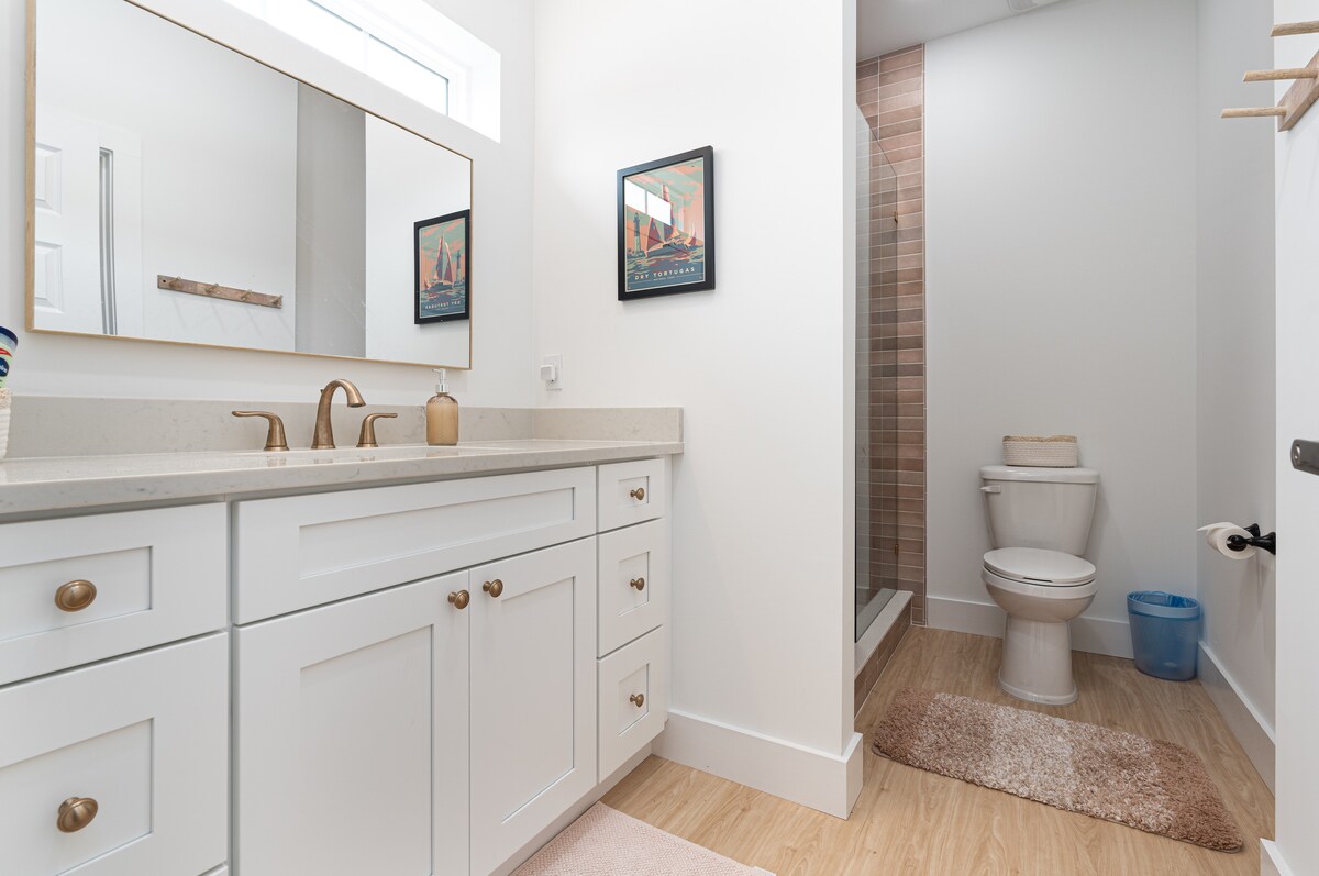 The main-floor en-suite: a long vanity, warm terracotta tile in the walk-in shower, and framed prints of the Florida coast on the wall. Everything within reach, and nothing shared with the rest of the house.