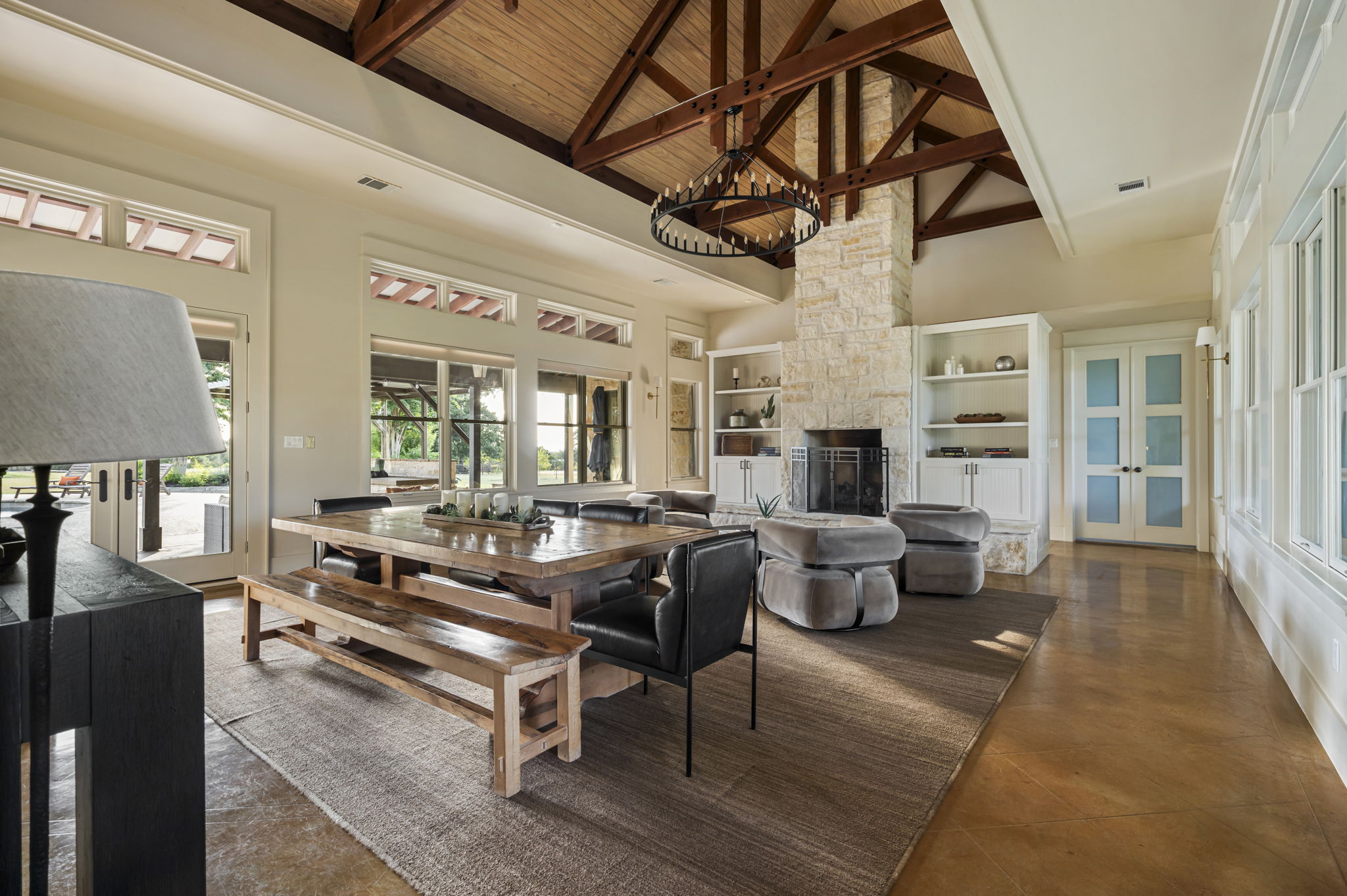A spacious and inviting living area featuring high ceilings with exposed beams, a large dining table with benches, a cozy fireplace, and ample natural light.