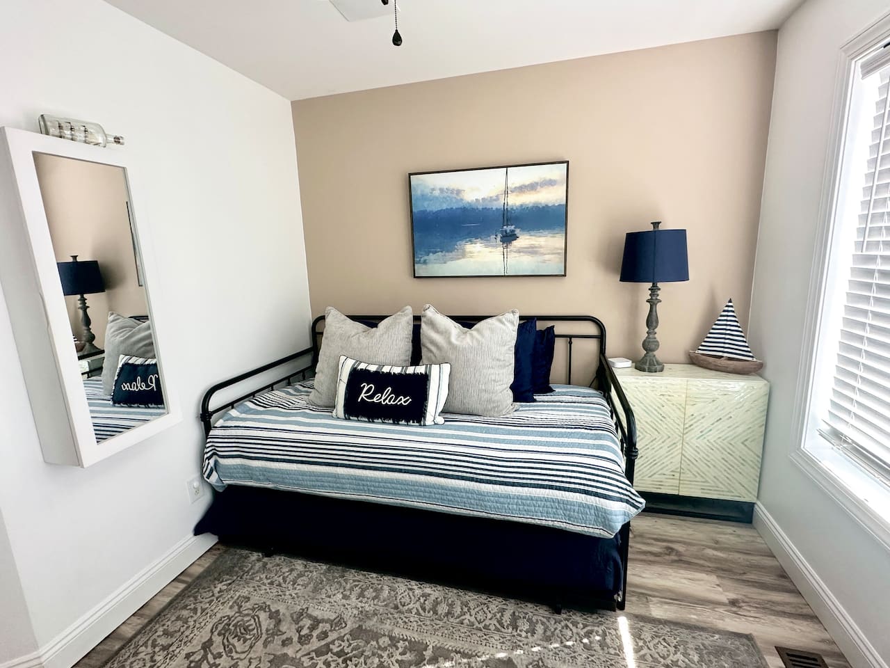This versatile sunroom features a black metal daybed with nautical pillows and a striped coverlet. The space includes a large patterned rug, a light-green storage cabinet with a sailboat decor piece, and a wall-mounted mirror.