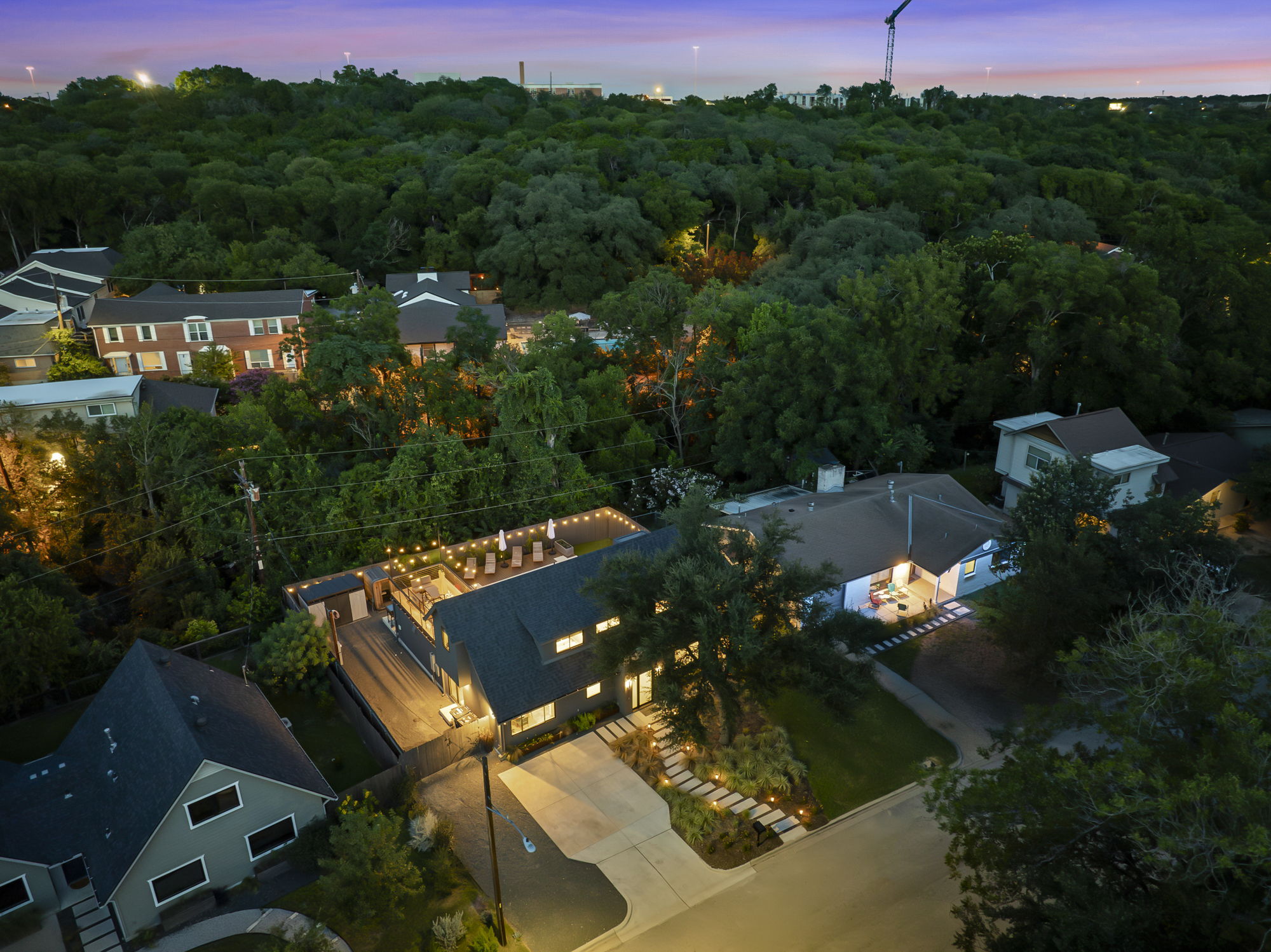 An aerial nighttime view of a modern home with a well-lit driveway and a spacious deck for entertaining, nestled within a lush, tree-filled neighborhood.