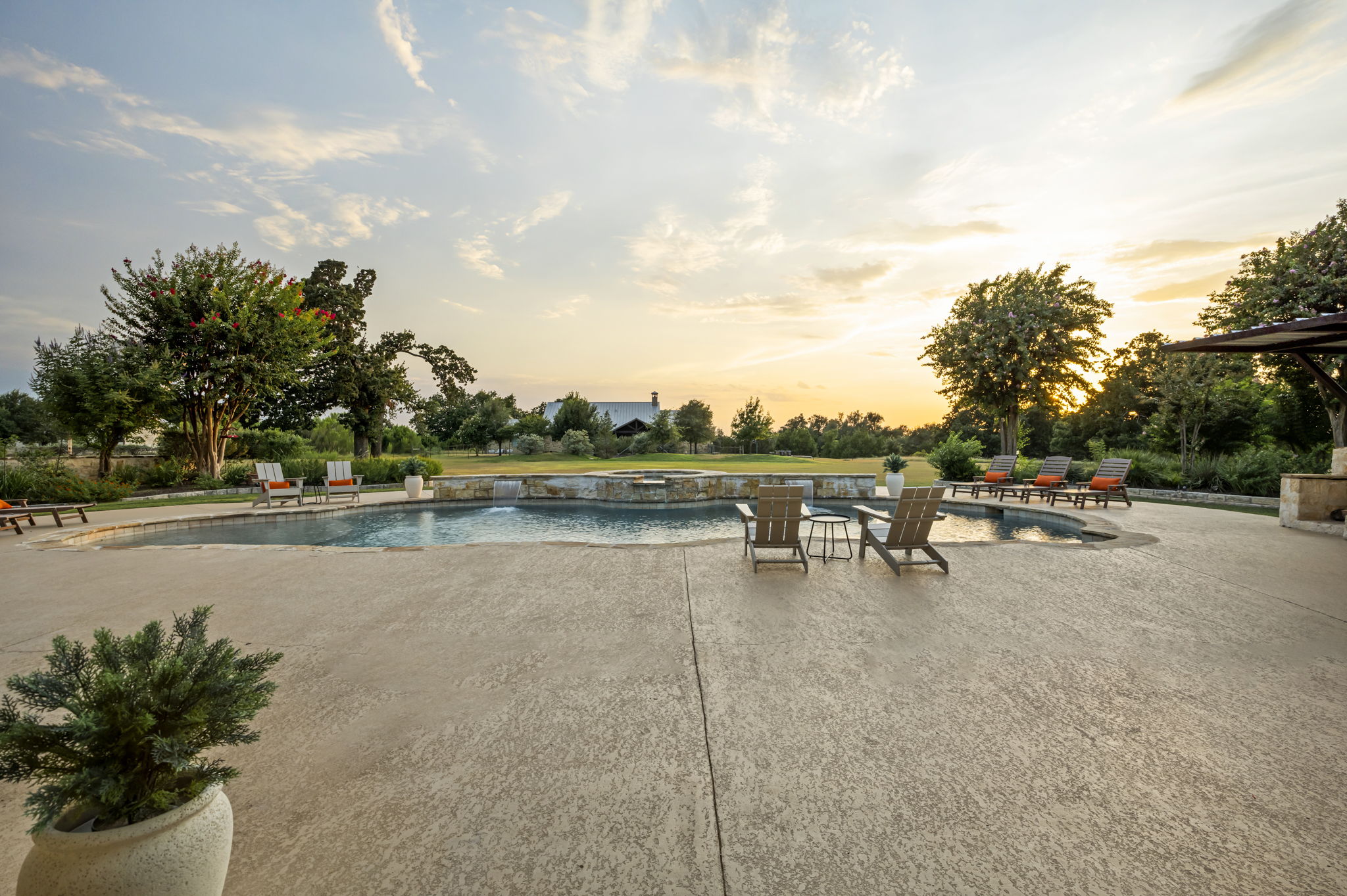 A luxurious Hill Country home featuring a sprawling outdoor living area with a resort-style pool, hot tub, and ample lounging space, set against a clear blue sky.