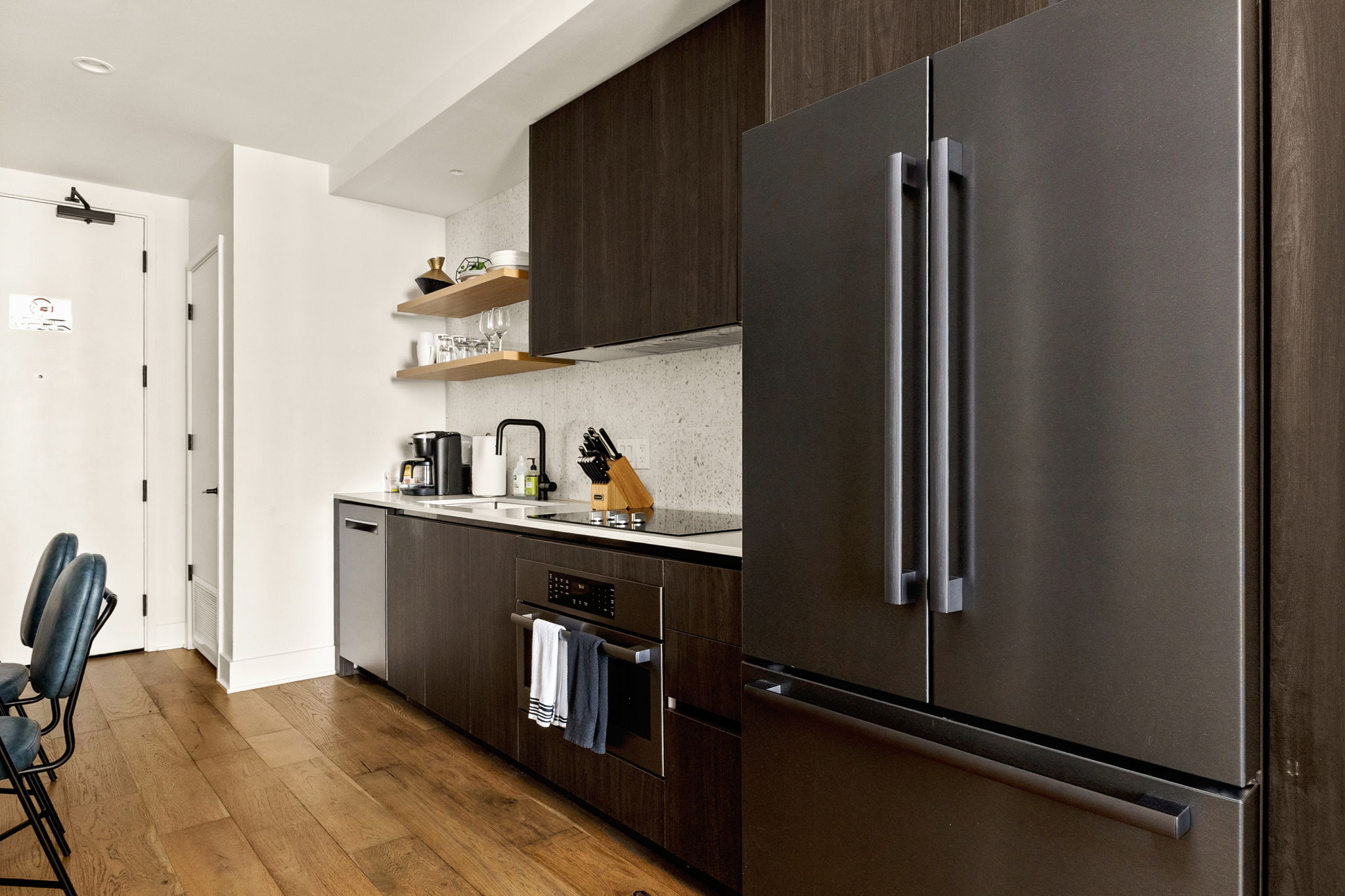 A modern kitchen featuring dark wood cabinetry, stainless steel appliances, and a light-colored speckled countertop with a matching backsplash. The space is equipped with a full-sized refrigerator, dishwasher, and stovetop, and is accented with open shelvi