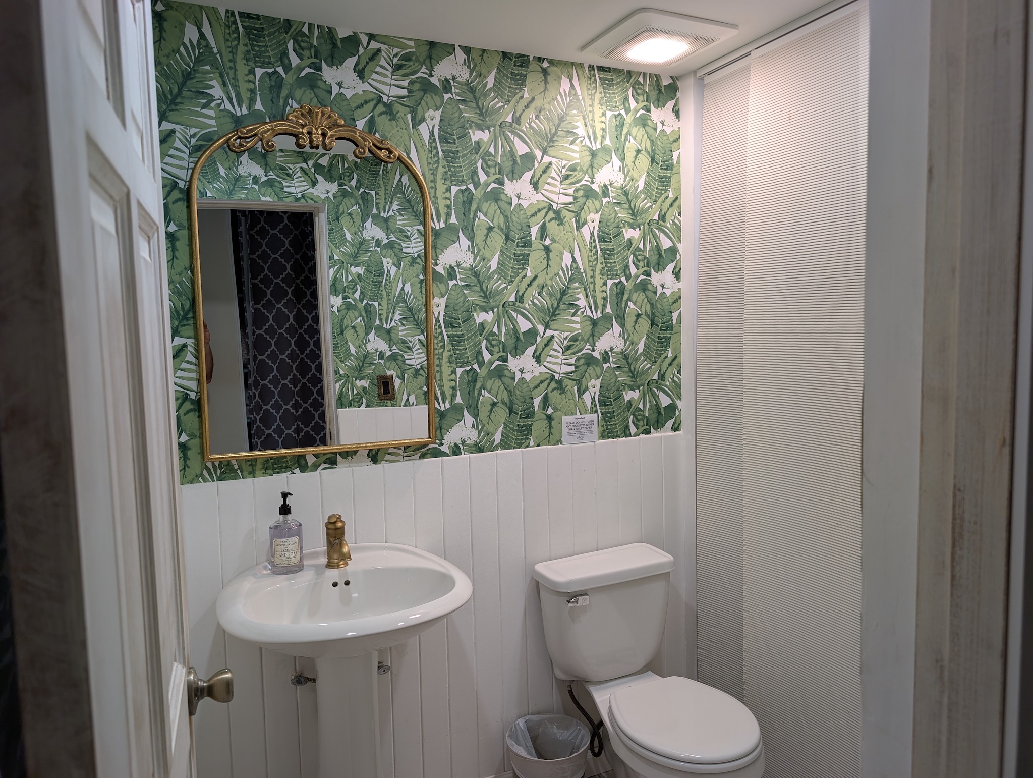 Social half-bath at Villa Coral featuring vibrant green tropical leaf wallpaper, white vertical shiplap wainscoting, a pedestal sink, and a vintage gold arched mirror in Dorado, Puerto Rico.
