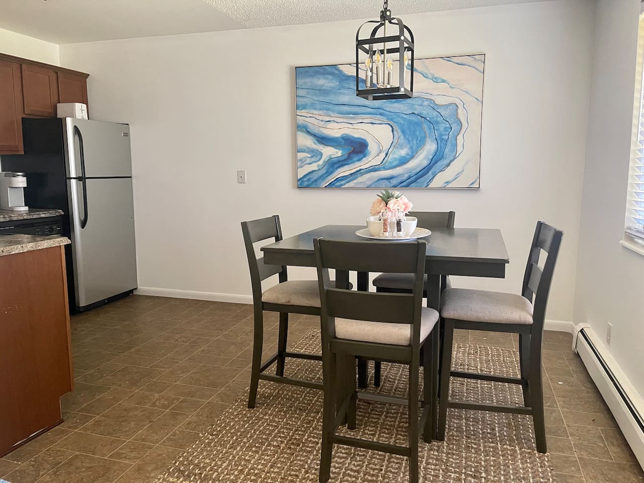 Bright, modern dining area with seating for four and eye-catching coastal artwork. Enjoy meals, game nights, or remote work under stylish lighting with natural light streaming in—an inviting space for connection and comfort during your stay.