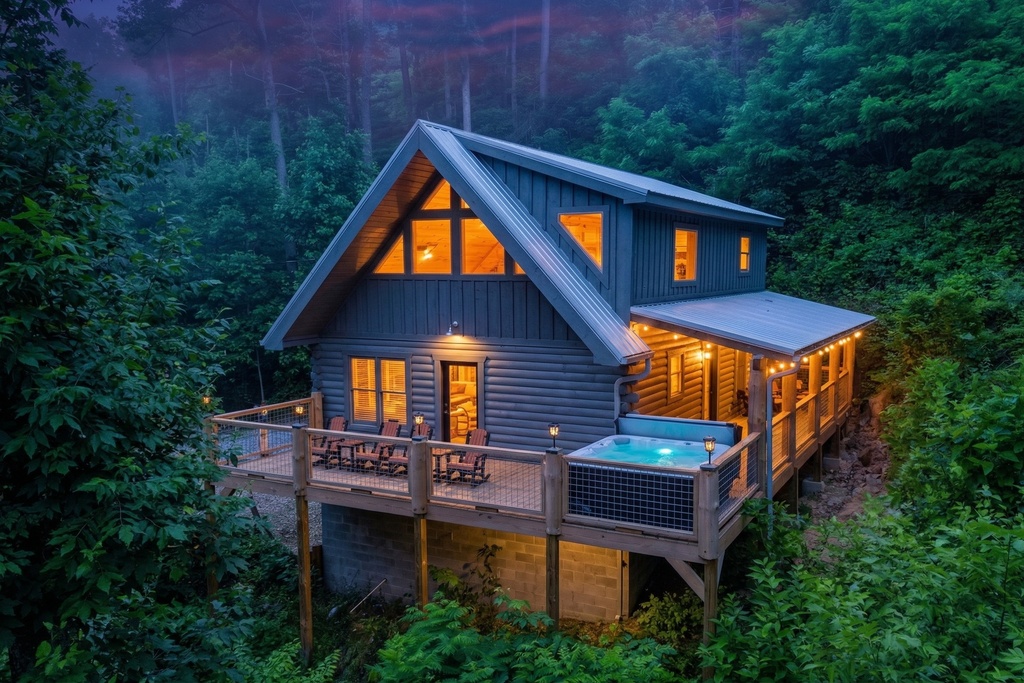 Bear Paws Cabin glows at twilight, nestled in the lush forests of Gatlinburg, Tennessee. This aerial view shows off the full property &mdash; spacious deck with rocking chairs, bubbling hot tub, covered porch with string lights, and soaring A-frame windows. The 