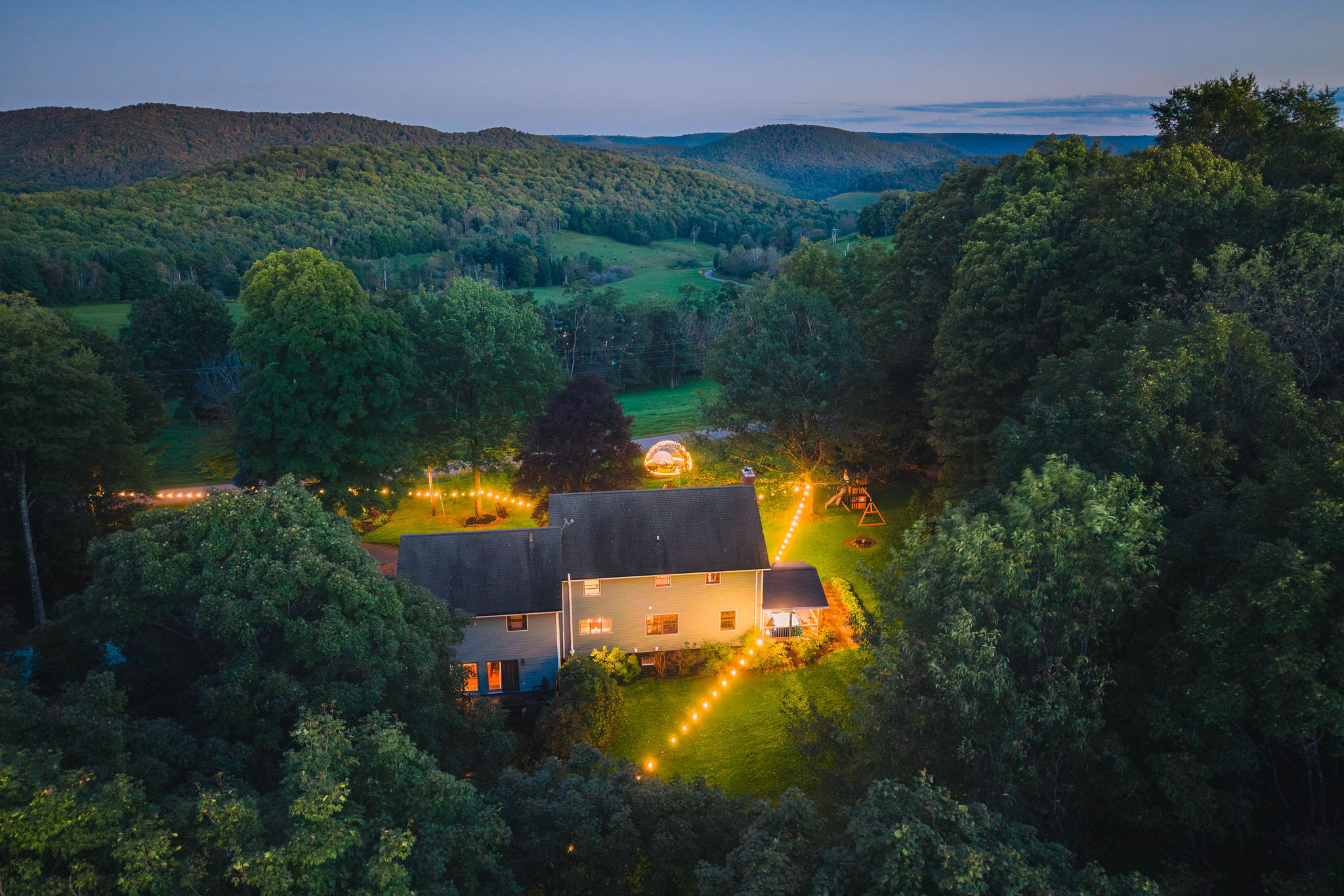 💡 The house lit up at dusk, dome and swing set on the lawn.