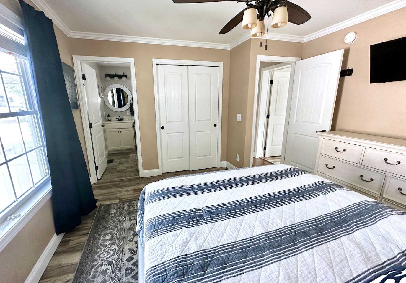 This bedroom provides the convenience of an en-suite bathroom and a large closet. The space is thoughtfully arranged with a dresser, wall-mounted TV, and hardwood-style flooring.