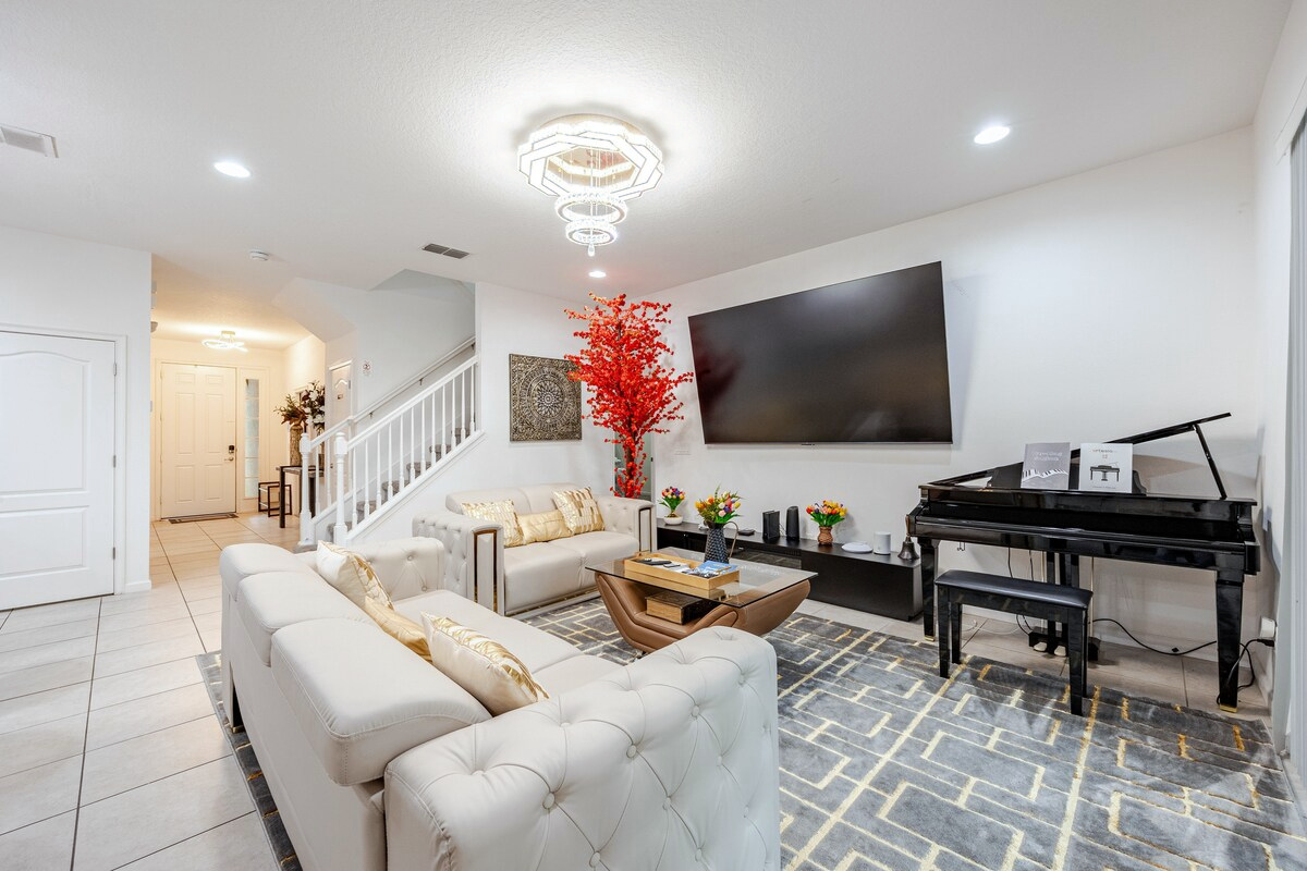 Main downstairs living room with a 98-inch TV, piano and electric fireplace, open to the kitchen and dining area.