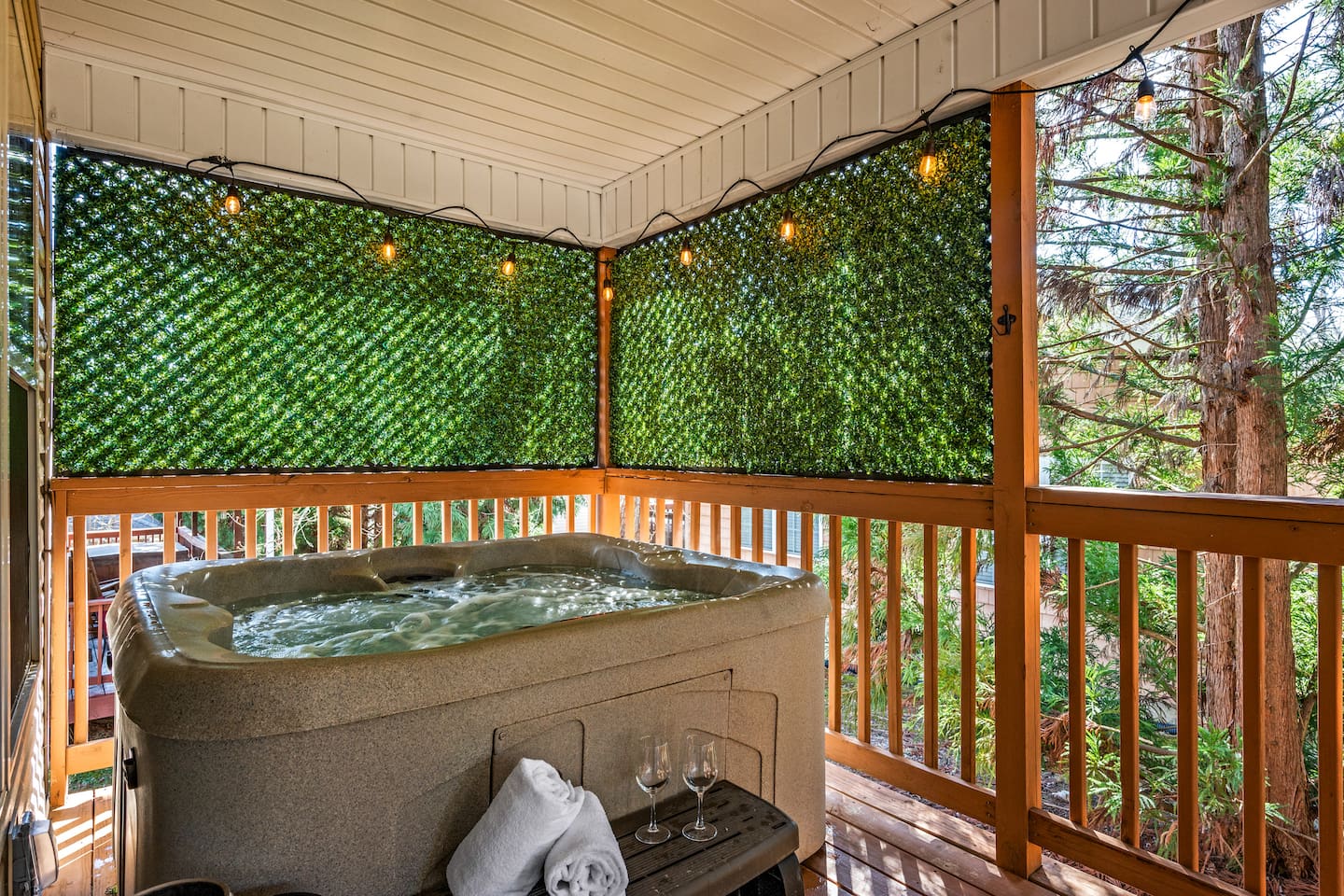This inviting covered deck features a private hot tub surrounded by lush greenery panels for added privacy. String lights create a cozy, ambient glow—perfect for unwinding in the evening. With soft towels, wine glasses, and peaceful wooded views, this spac