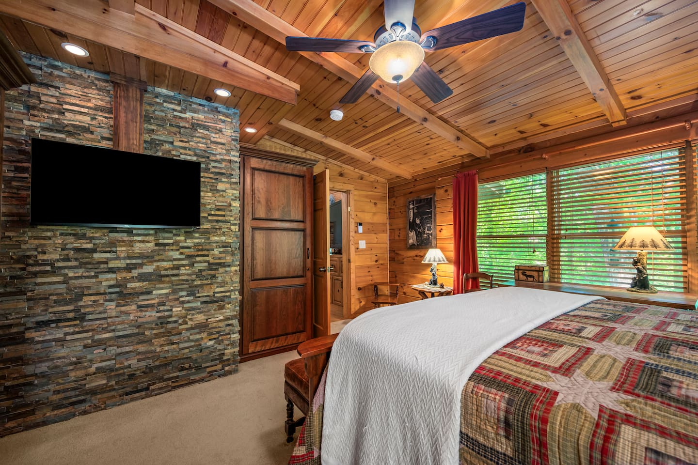 Master bedroom includes a comfortable, queen bed with vaulted ceilings, a stunning stone accent wall with mounted TV. Large windows bring in natural light and peaceful wooded views, creating the perfect space to relax and unwind after a busy day.