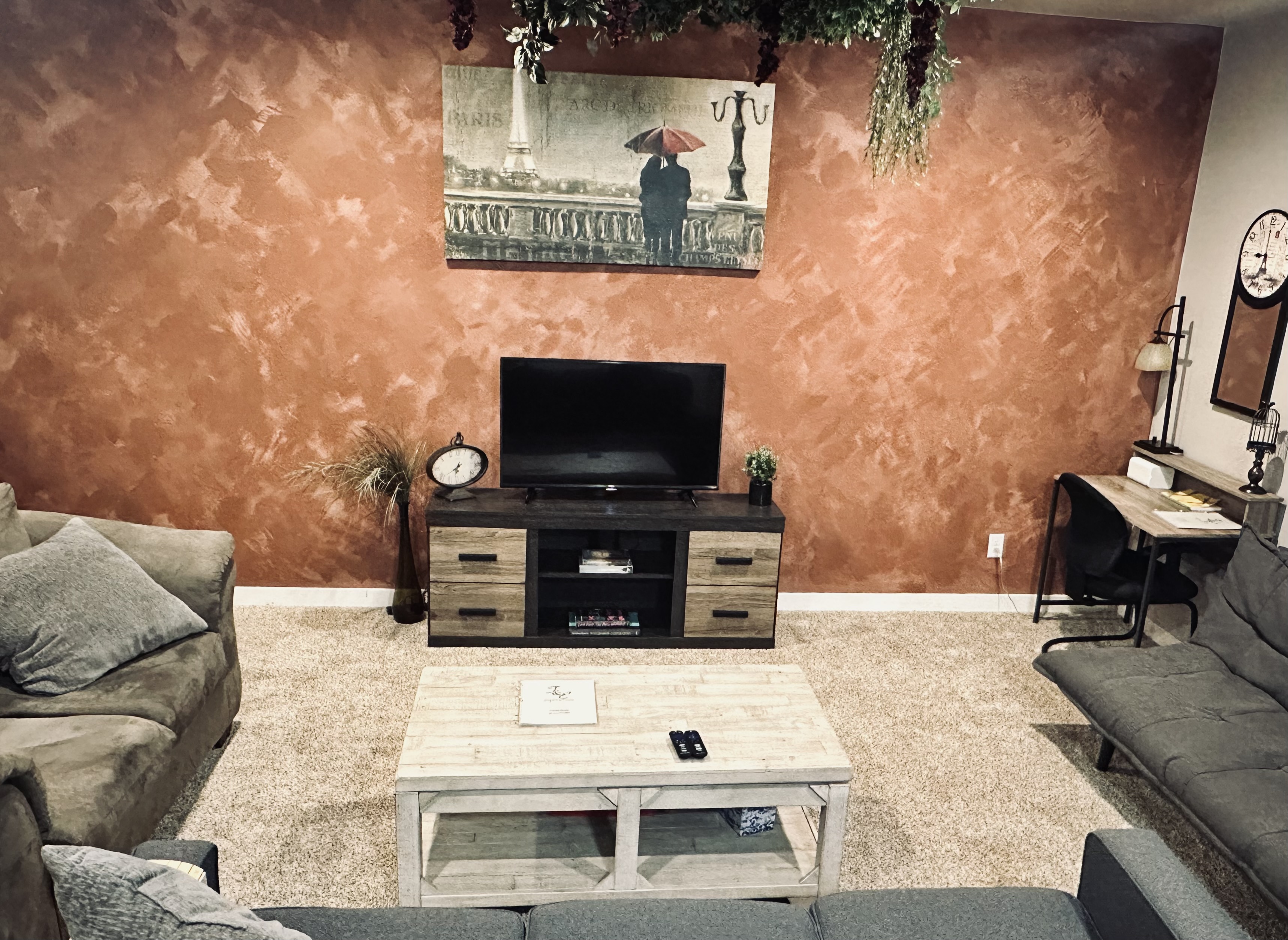 A cozy living room features a textured orange wall, a large painting of a couple under an umbrella in Paris, a flat-screen TV on a dual-tone wooden cabinet, a small clock, potted plants, and a light-colored wooden coffee table with remote controls on top, alongside a desk with a chair and a mirror in the corner.