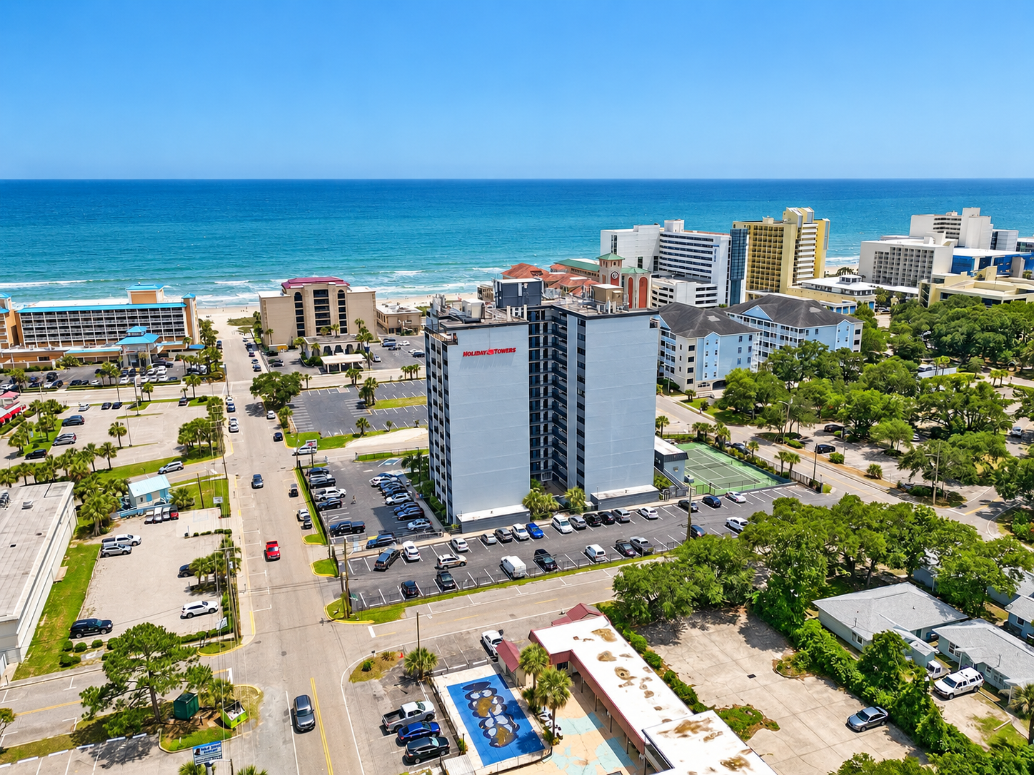 Aerial view of Holiday Towers with shared pool, hot tub, sports court, parking, and Myrtle Beach attractions nearby.