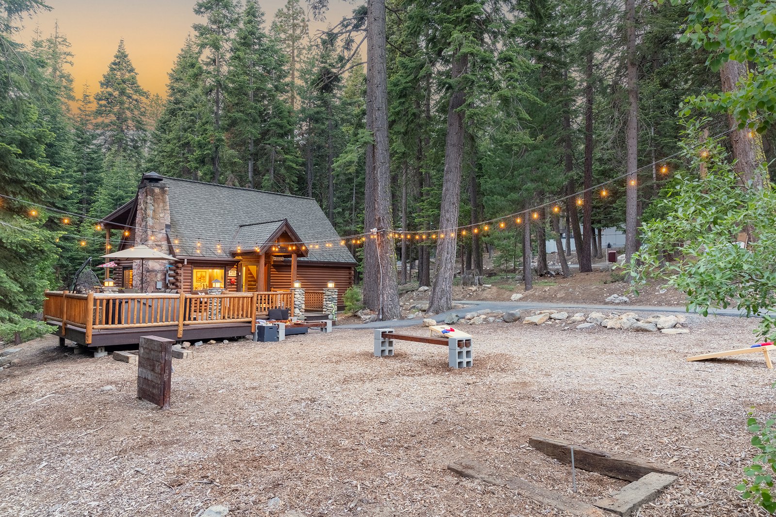 Step out back as the sky turns pink — cornhole set up, string lights flickering on, the cabin glowing through the firs.