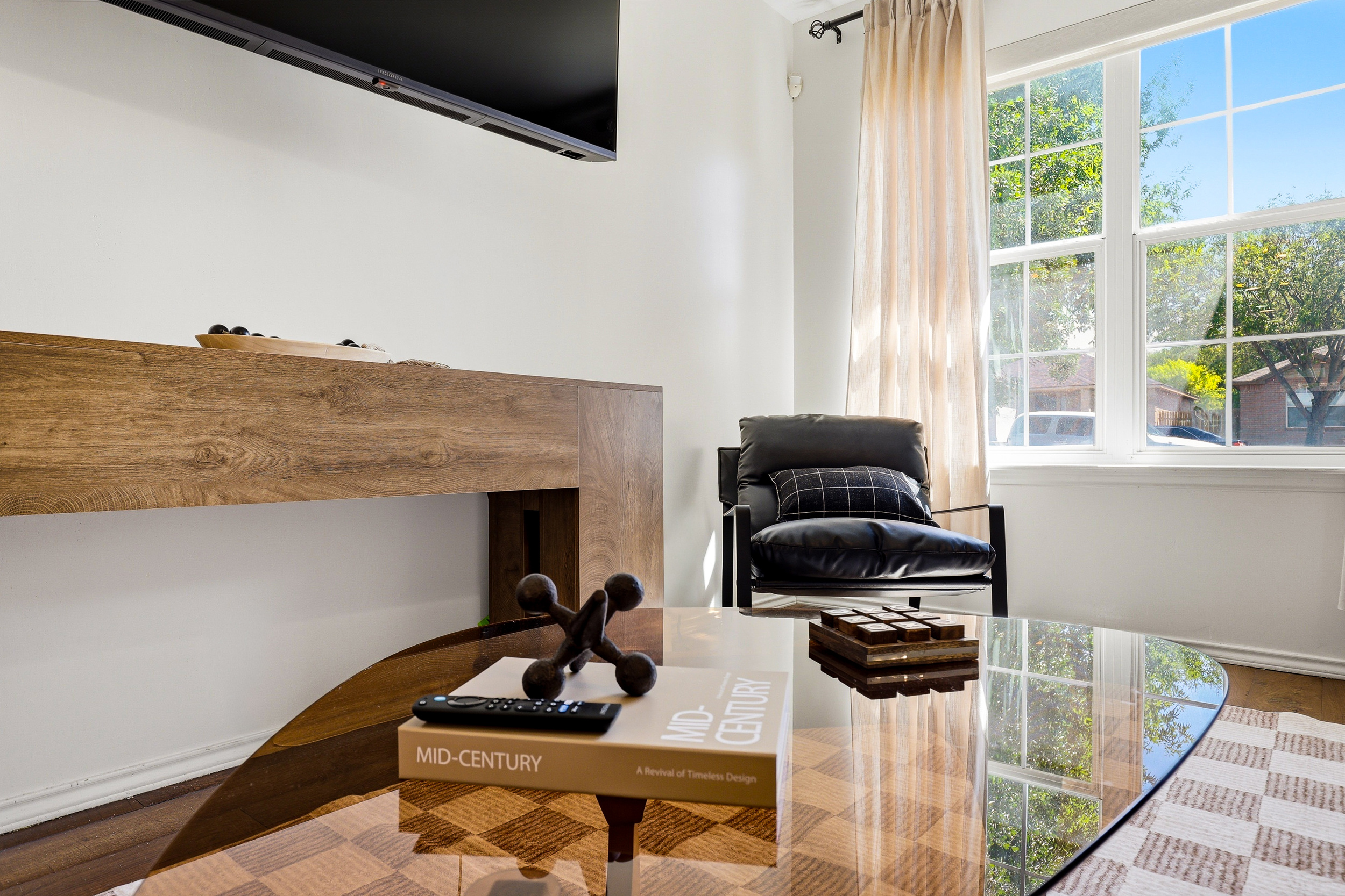 Living room detail showing wall-mounted Smart TV, wood console, leather accent chair, and large picture window with natural light. Bright and airy with a mid-century feel throughout.