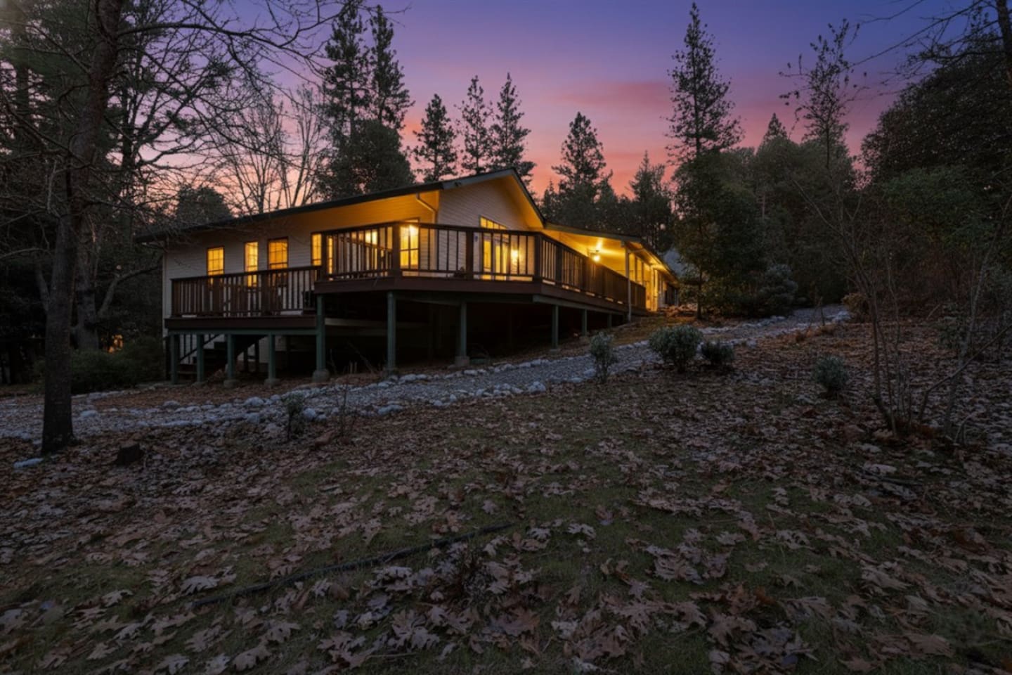 Cabin exterior at sunset with a wraparound deck, wooded setting, pine tree surroundings, and warm evening lighting for a peaceful mountain retreat.