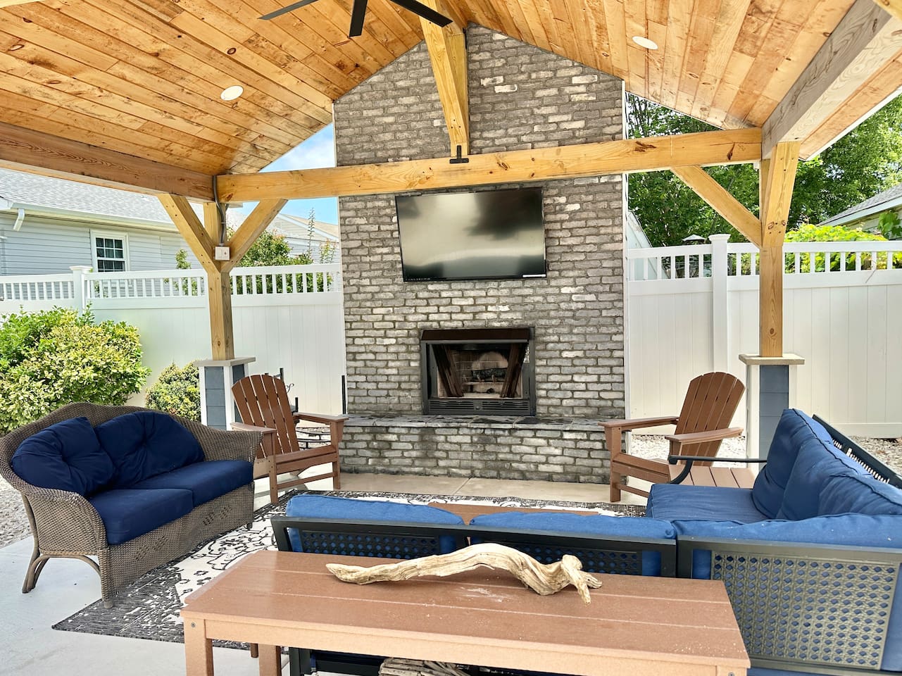 This shaded pavilion features a brick fireplace and a large flat-screen TV for outdoor entertainment. The seating area includes navy blue sofas and wooden Adirondack chairs under a vaulted ceiling.