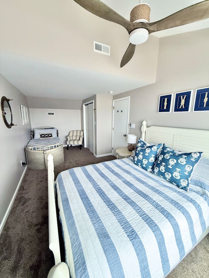 This family-friendly bedroom features a white bed dressed in a blue and white striped quilt, accented by decorative patterned pillows. The space also includes a charming, curved boat-style bed perfect for children.