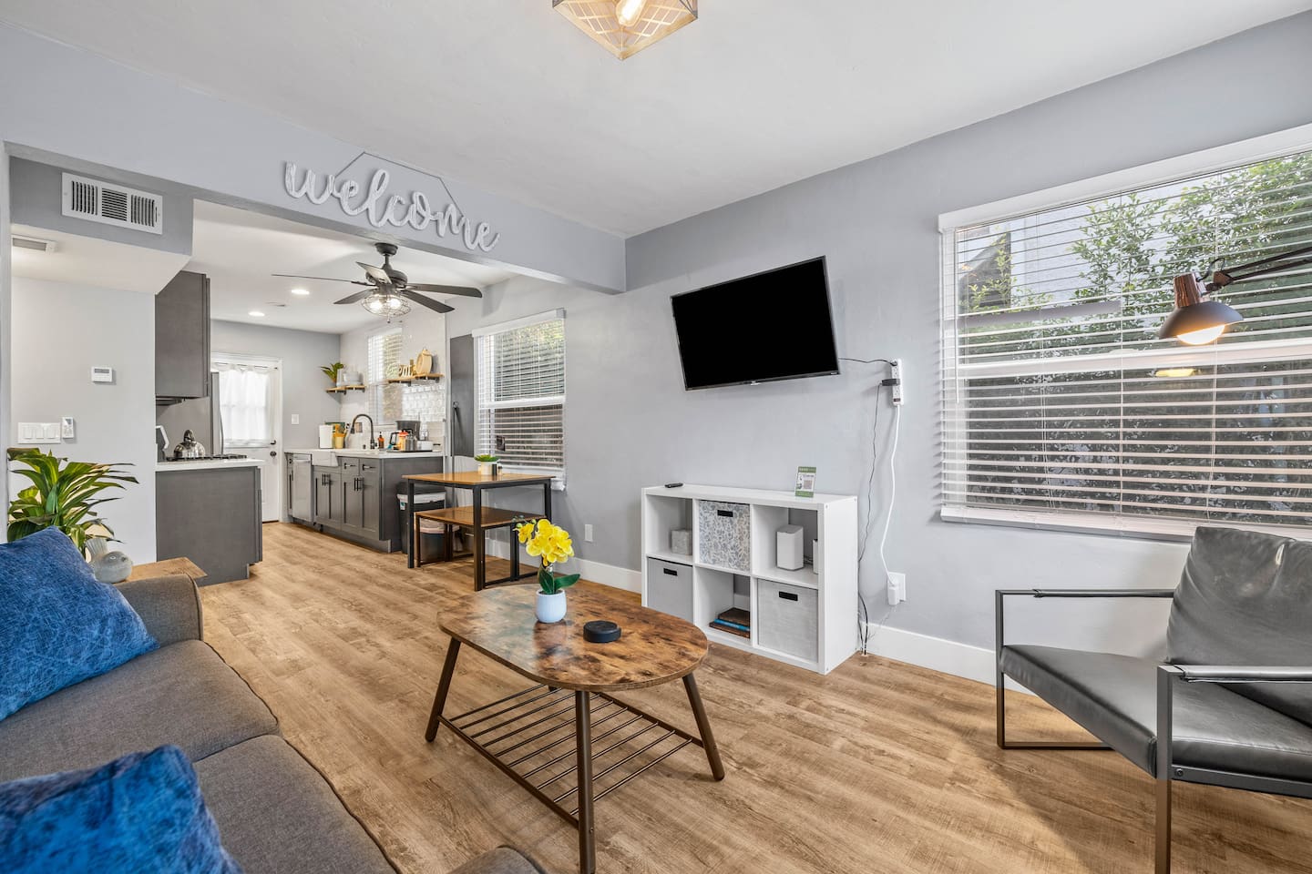 Experience comfort in our aesthetic living room, featuring a charming sofa, and a decorative coffee table. A welcoming sign sets the tone for relaxation, with a TV and softly lit chair for your enjoyment.