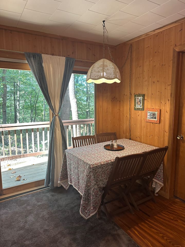 The dining area features a leaf table with four chairs, plus two extras stored in the cabin. It sits in front of a sliding glass door, offering a nice view and natural light. Great for meals, puzzles, or planning your day in the orchard.