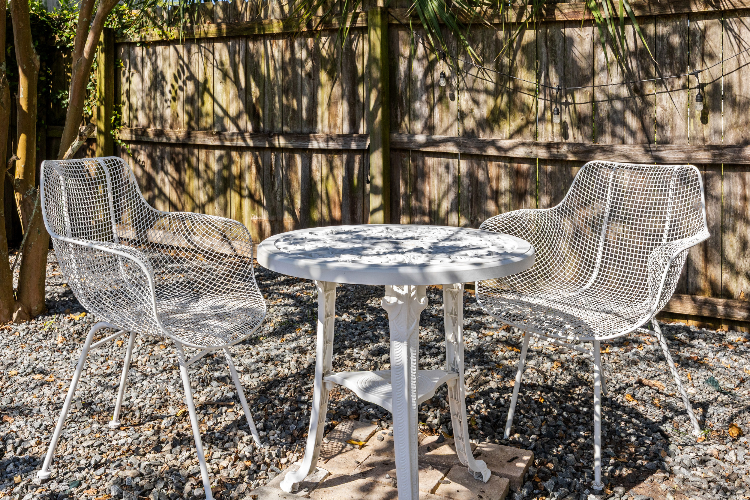 Cozy Bistro Seating
A quaint outdoor seating nook with white metal chairs and a matching table, set against a wooden fence with string lights. A peaceful spot for morning coffee or evening conversation.