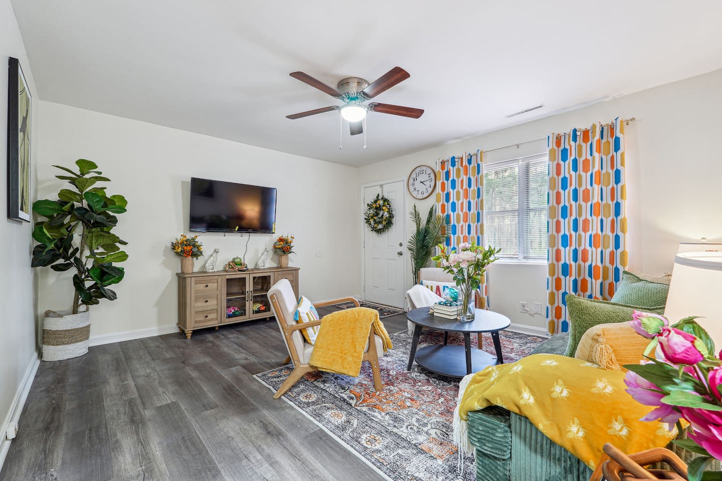 Enjoy a cozy living room featuring a long comfortable sofa, a TV for your entertainment, a ceiling fan, two chair, and a window with a lovely view outside — perfect for relaxing and unwinding.
