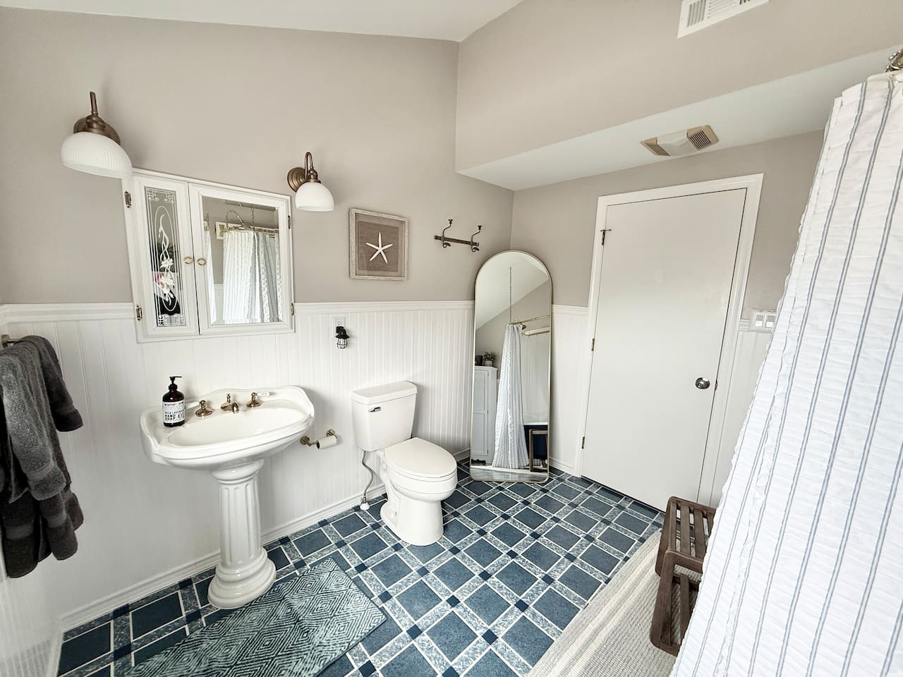 This spacious coastal-style bathroom features a white pedestal sink, blue and white checkered tile flooring, and nautical decor like a starfish and arched mirror.