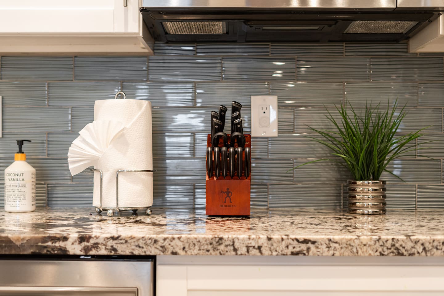 Close-up of a modern kitchen counter with granite surfaces, a glossy tile backsplash, and neatly arranged essentials, including a knife block and greenery.