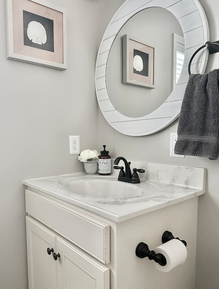This bathroom vanity features a marbled white countertop with a recessed oval sink and a dark, two-handle faucet. The white cabinet base has dark knobs, and a matching dark toilet paper holder is mounted to its side.