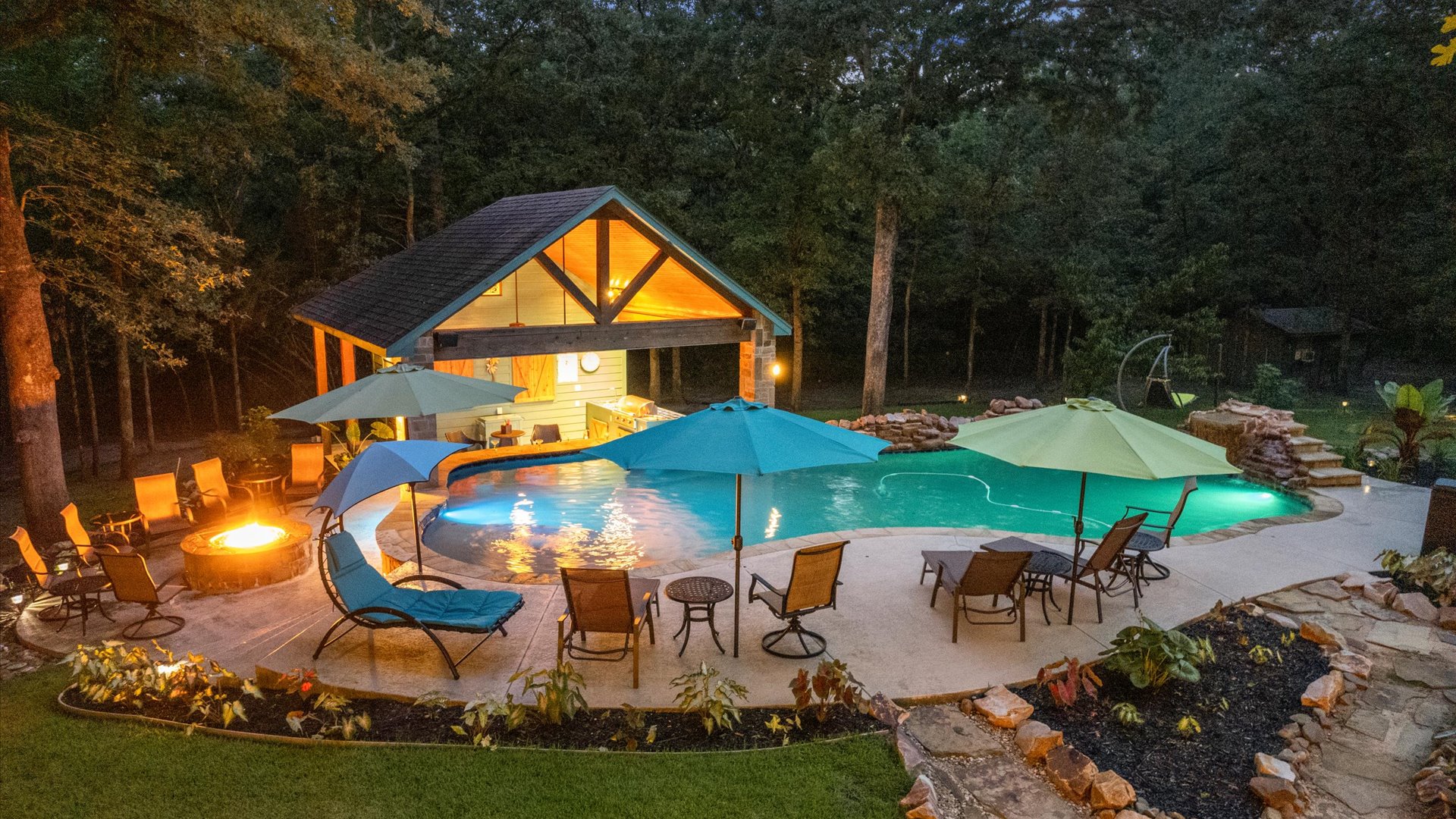 A stunning backyard oasis at night! Relax by the glowing fire pit, lounge by the pool, or enjoy the cozy ambiance of the illuminated outdoor kitchen and swim-up bar—perfect for unwinding under the stars