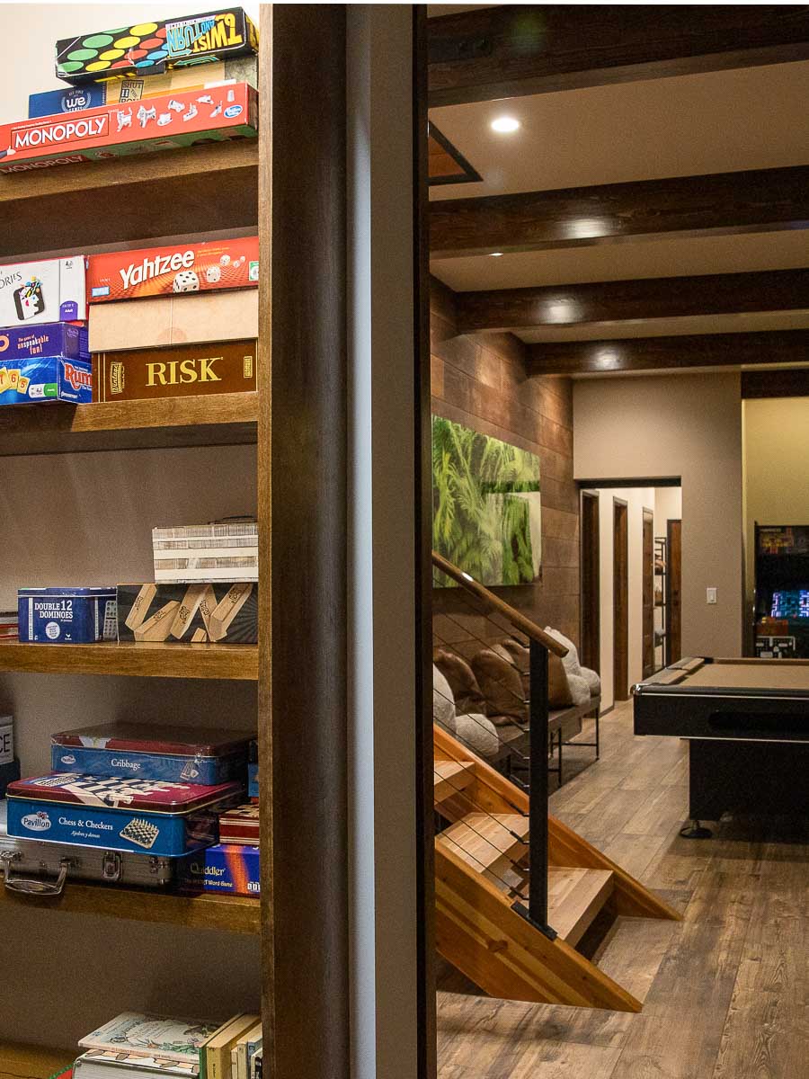 The Library Nook at Sunset Falls &mdash; books and board games on built-in shelving, beam ceiling, stairway descent, and through-view to pool table and arcade beyond