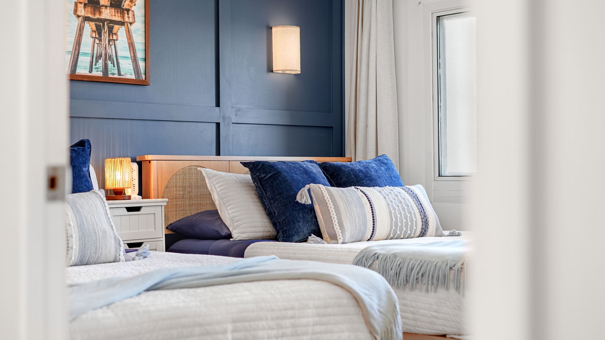 The bedroom features 2 queen beds, storage baskets for throw pillows and blankets, cordless sconces, blackout curtains, luxury bedding and stunning beach view from the panoramic window