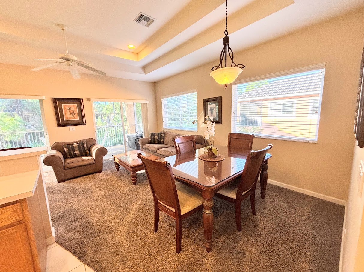 Bright open dining and living area with tray ceilings, comfortable seating, large windows, and sliding glass doors leading to the private balcony.