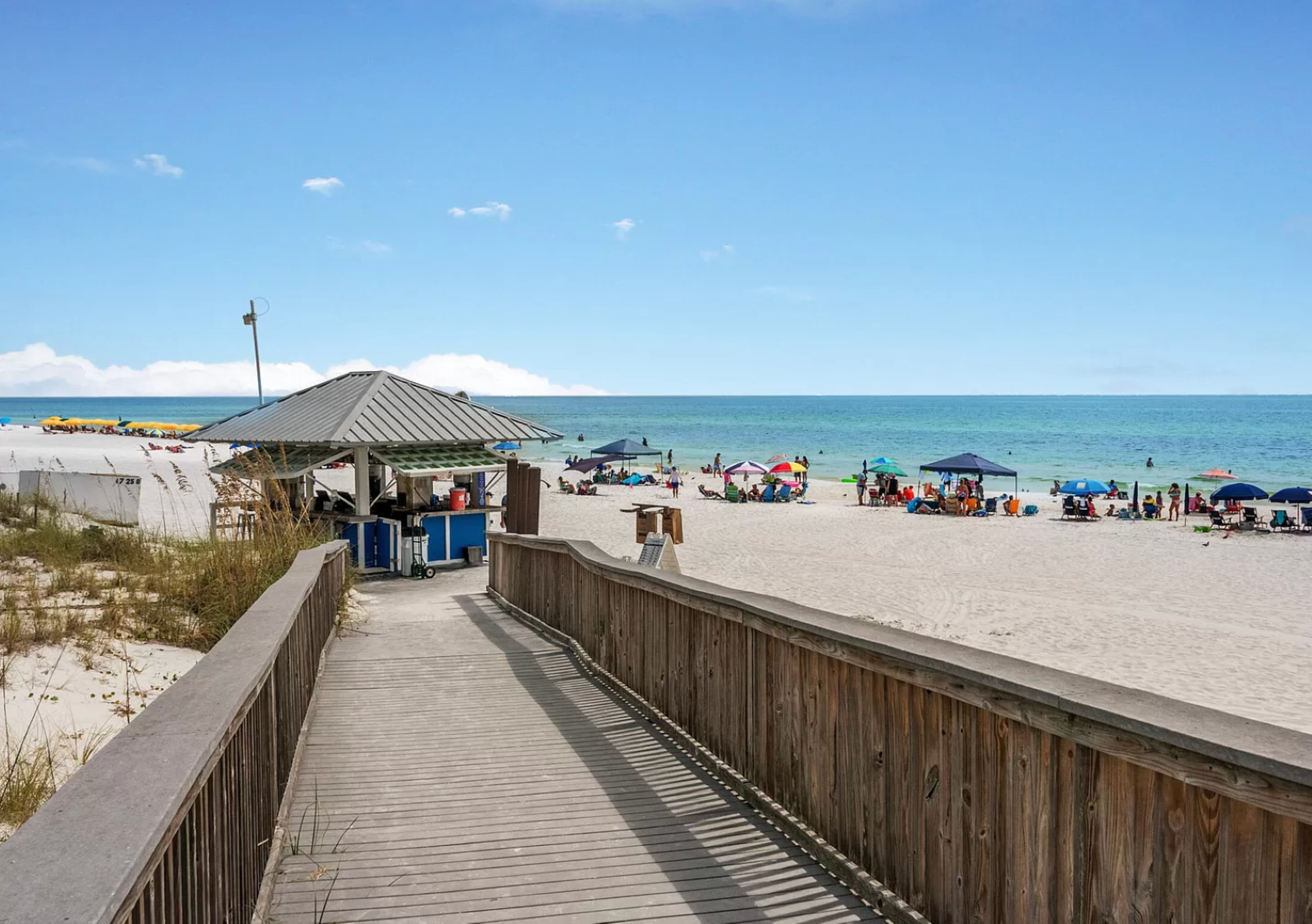 A wooden boardwalk leads to the shore. Gentle waves, colorful umbrellas, and bathers create a lively, family-friendly coastal atmosphere.