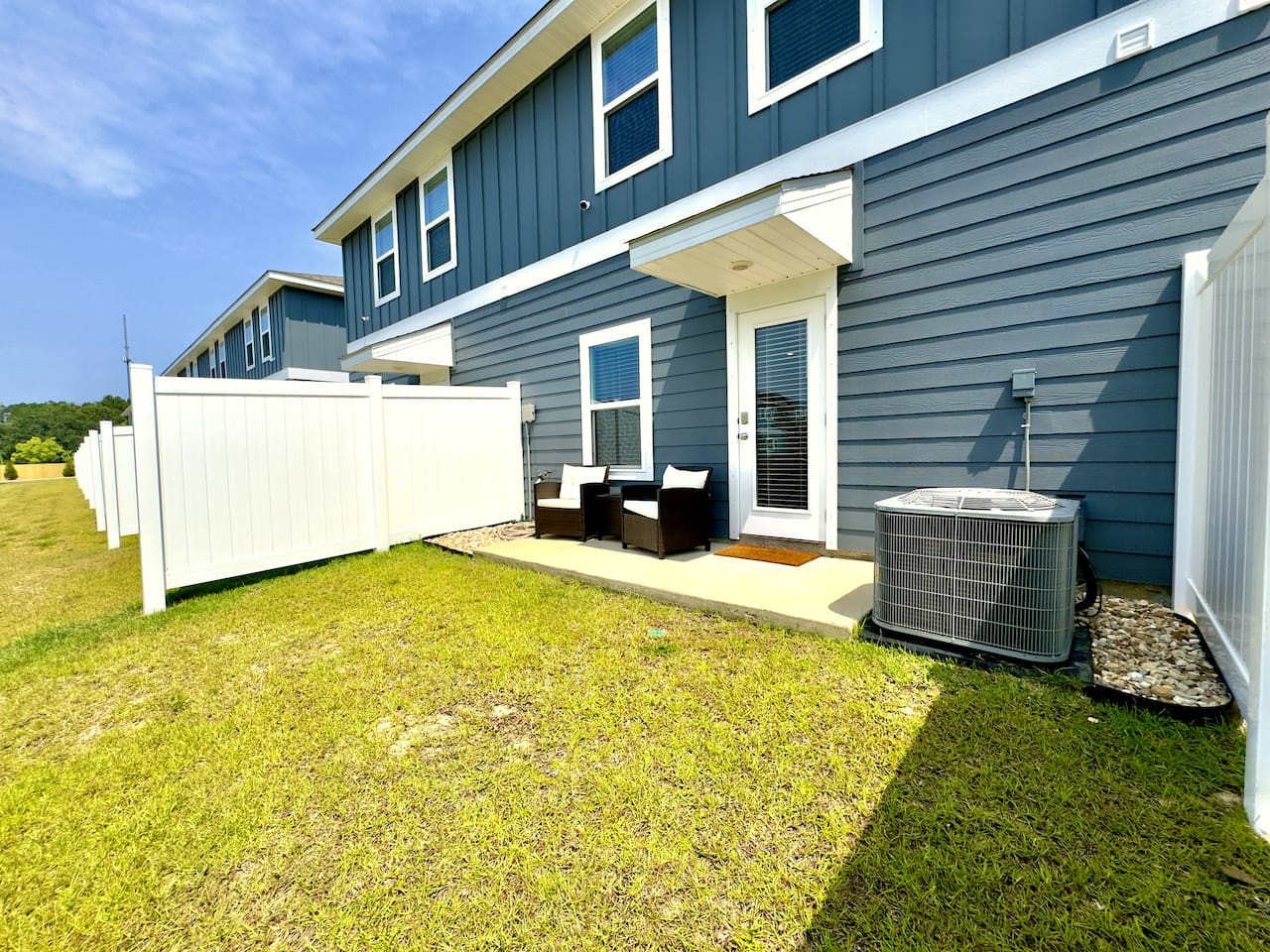 Stepping outside, soft grass underfoot, warm sun overhead. The private patio invites a cold beer, good conversation, or a grill session with friends. Coastal blue siding, fresh air, easy and open.