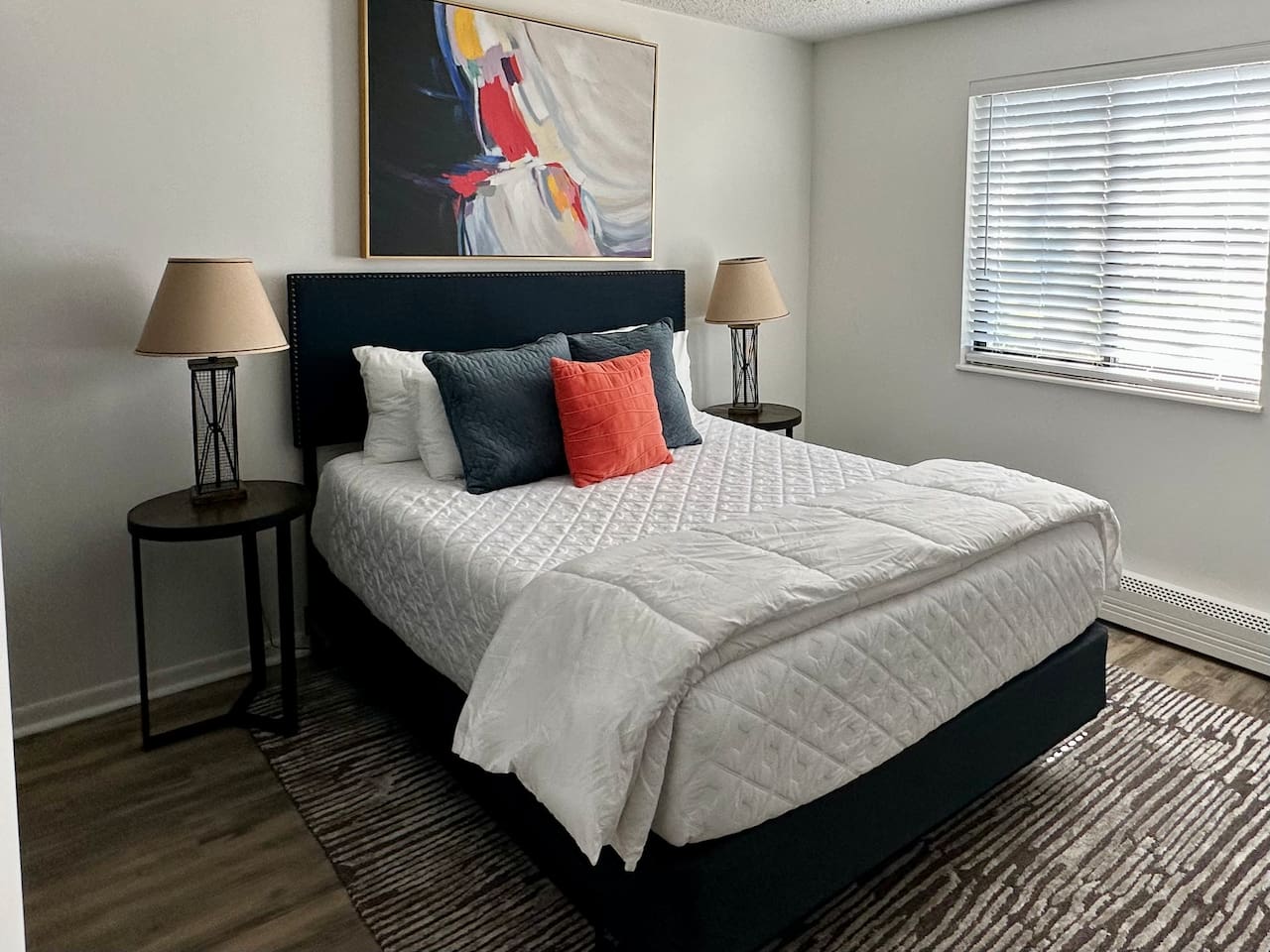 Rest easy in this bright, modern bedroom featuring a plush queen bed with cozy linens and colorful accents. A large mirror, natural light, and calming decor create a peaceful retreat—perfect for a great night’s sleep during your short-term stay.