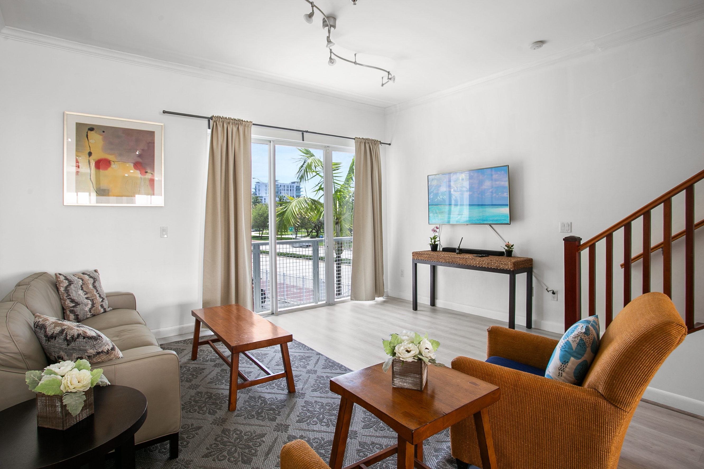 Living room with leather sofa, accent chairs, and a 50-inch flat screen TV. Sliding glass doors open to a Juliet balcony for sea breeze.