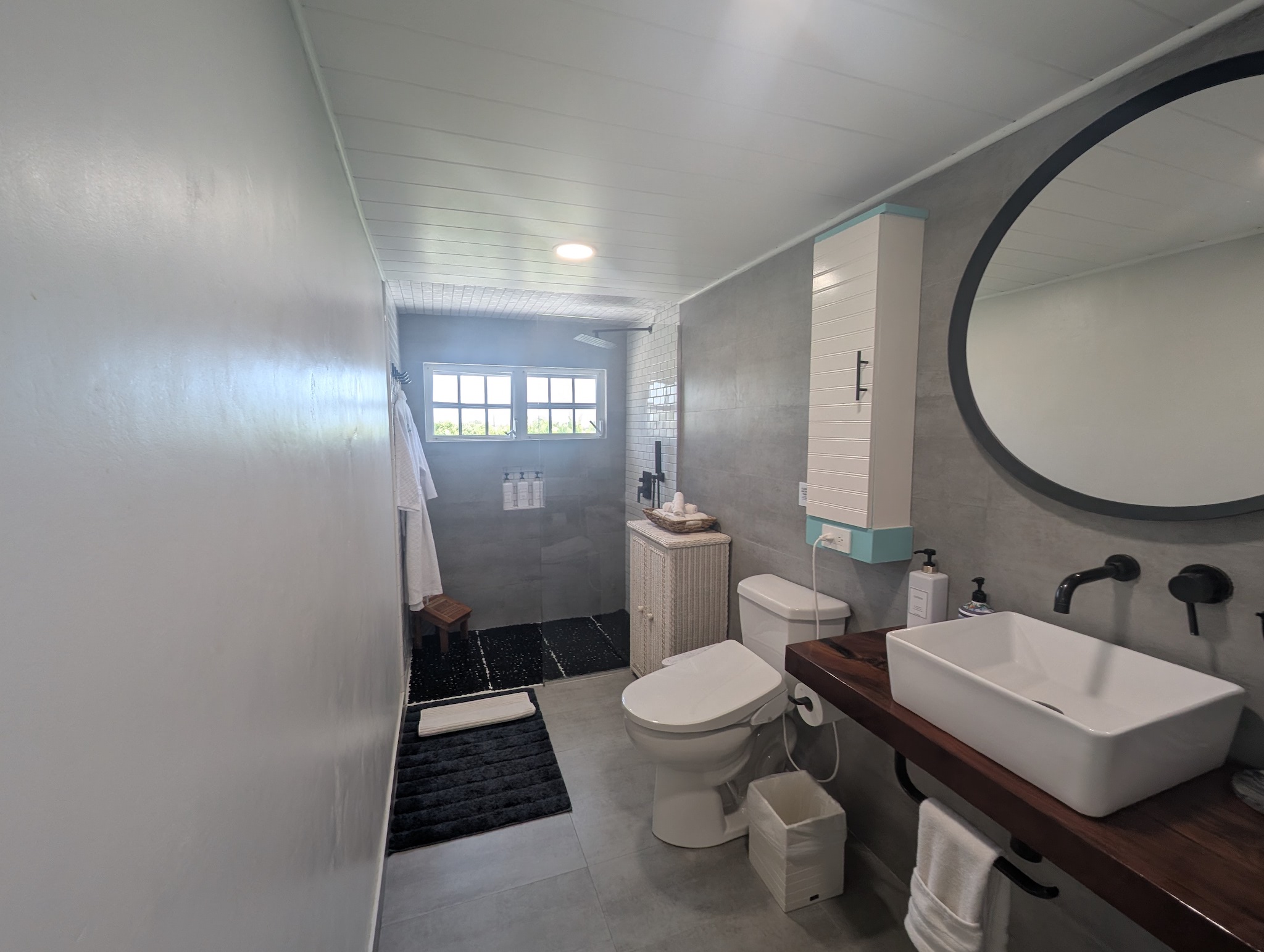 Modern upstairs bathroom at Villa Coral featuring a wood vanity with a white vessel sink, matte black wall-mounted faucets, large round mirror, concrete-gray tiles, and a walk-in shower in Dorado, Puerto Rico.
