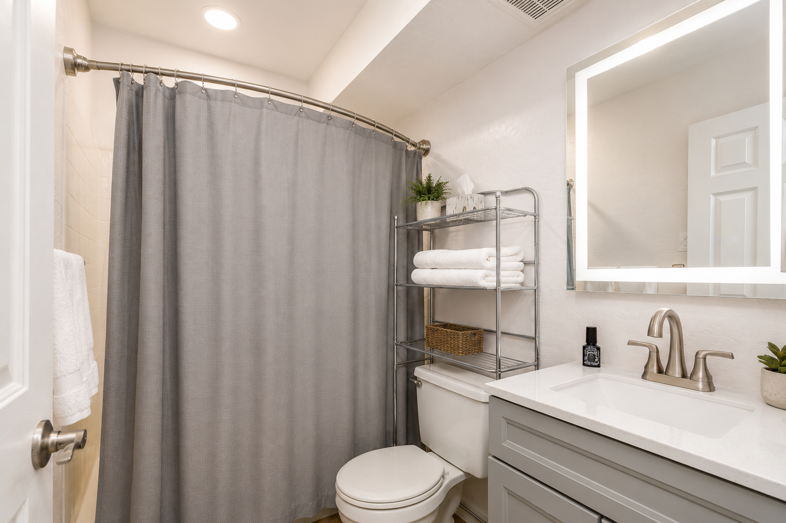 Full bathroom with gray vanity, lit mirror, shower/tub combo, and shelf storage for towels and bath items.
