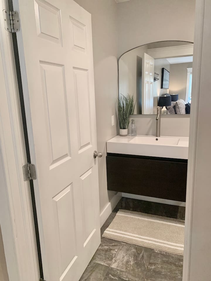 This sleek half-bath features a contemporary floating vanity with a clean white countertop and a minimalist faucet. The modern design is accented by a simple rug and a touch of greenery, providing a stylish and convenient space for guests.