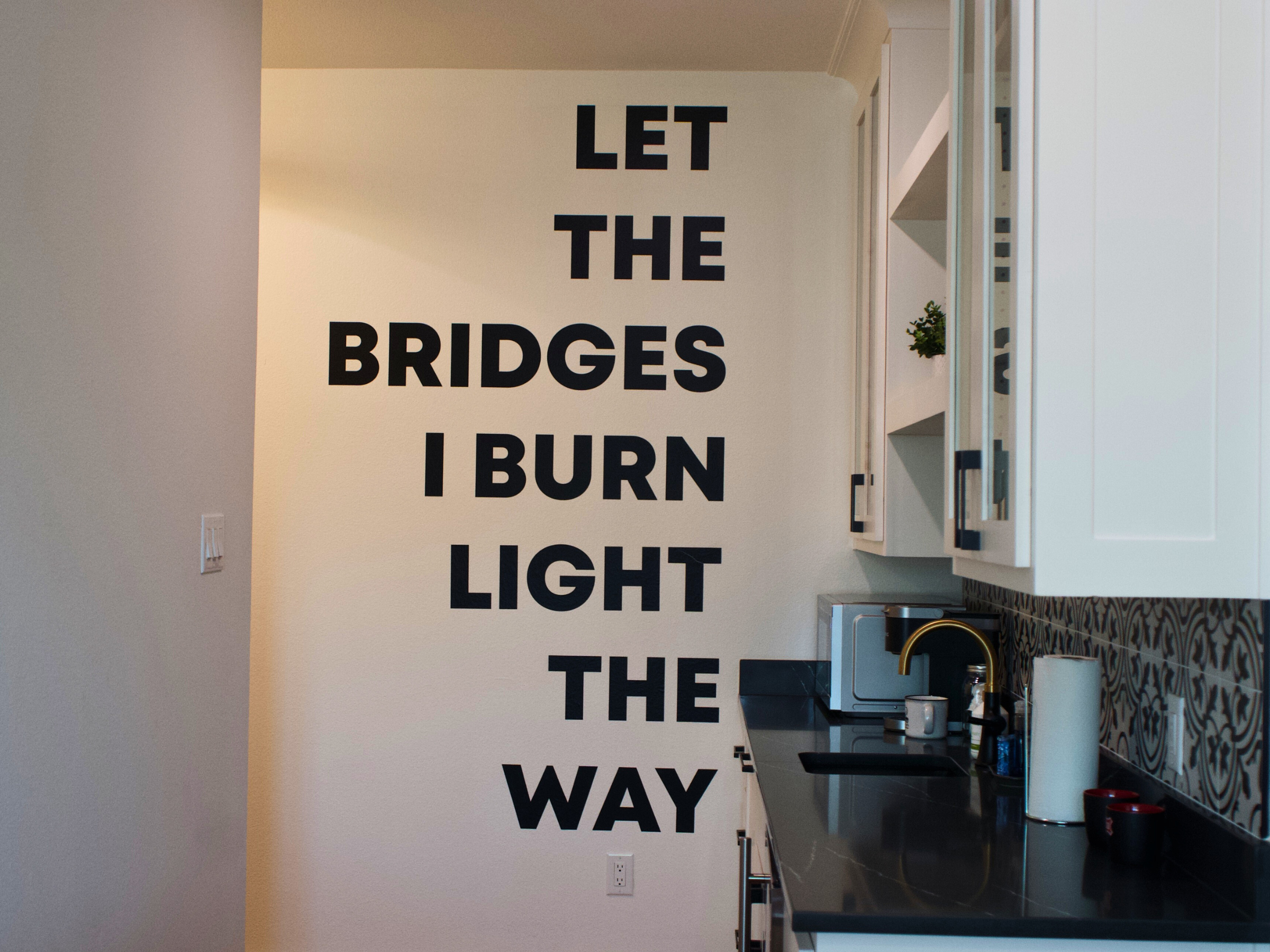 Bold wall art reading “Let the bridges I burn light the way” adds a modern, confident statement to the kitchen, giving the space a unique, urban character.